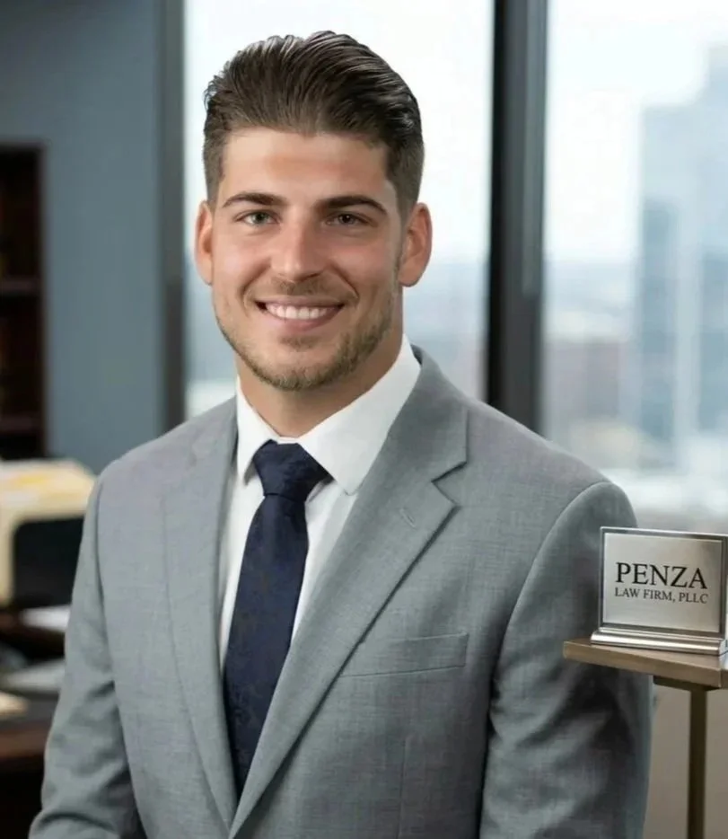 A young man with neatly styled brown hair, a trimmed beard, wearing a light gray suit, white shirt, and navy blue tie, smiling against a black background.