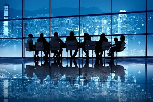A group of people sitting at a conference table on a high floor of a building, with a city skyline and mountains visible through large windows behind them.