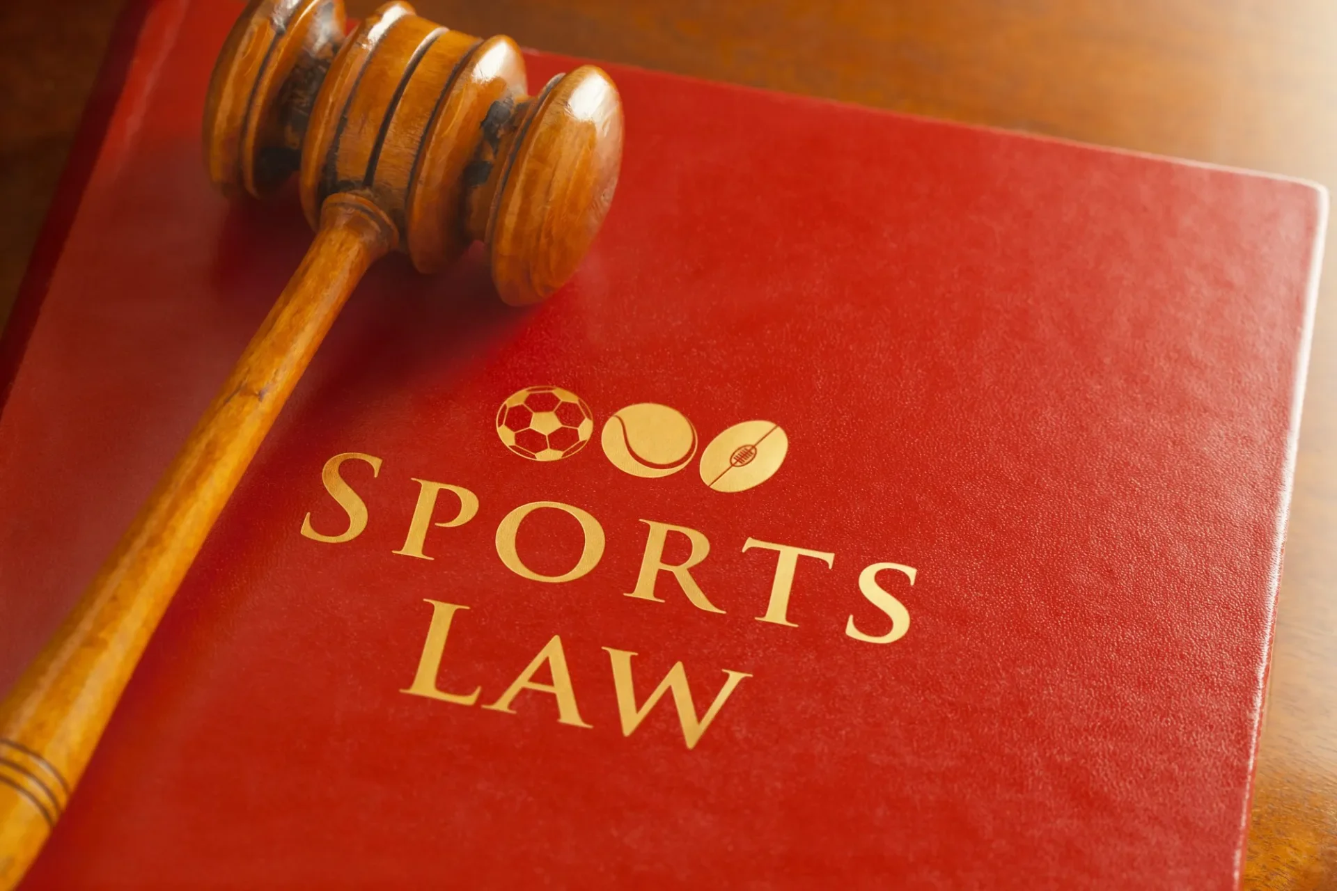 A wooden gavel resting on a red book titled 'Sports Law' with icons of a soccer ball, tennis ball, and baseball above the title.