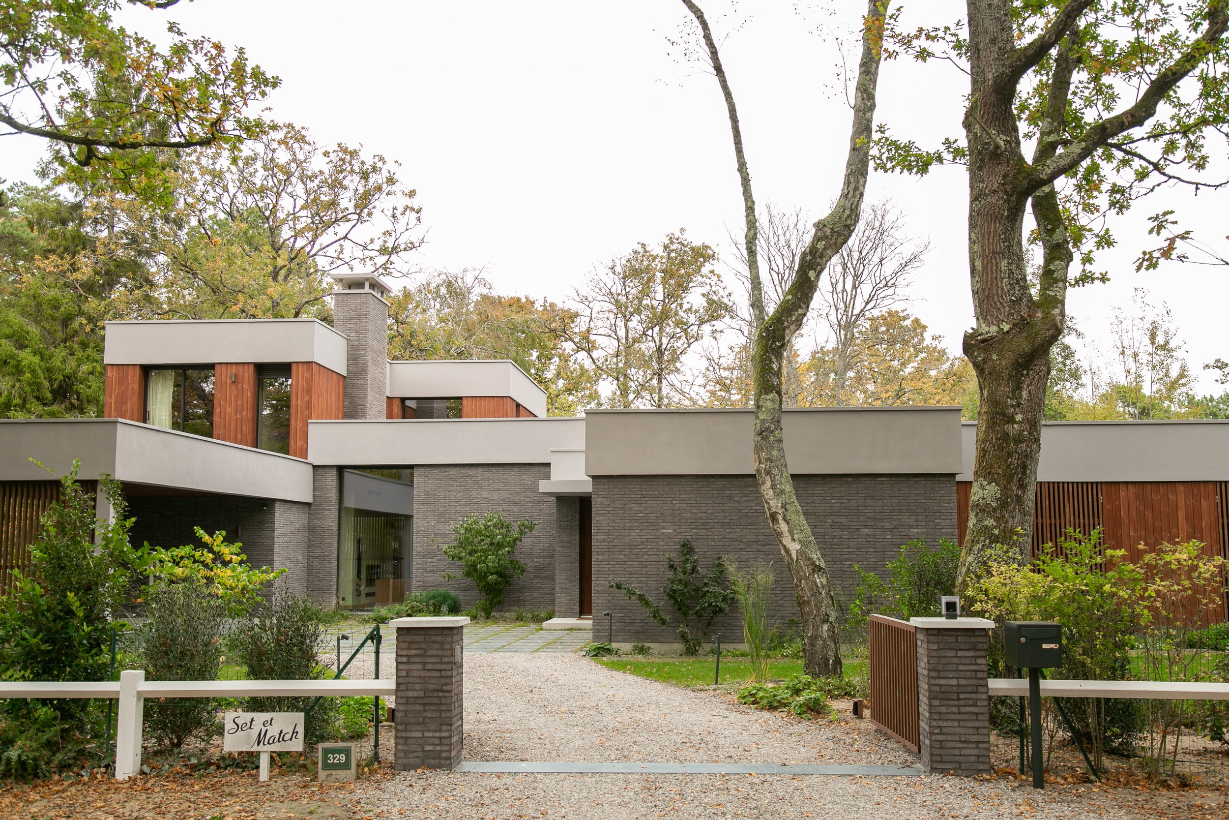 Maison moderne en pierre et bois, entourée d'arbres, avec un chemin en gravier à l'avant et une clôture en bois, dans un environnement résidentiel.