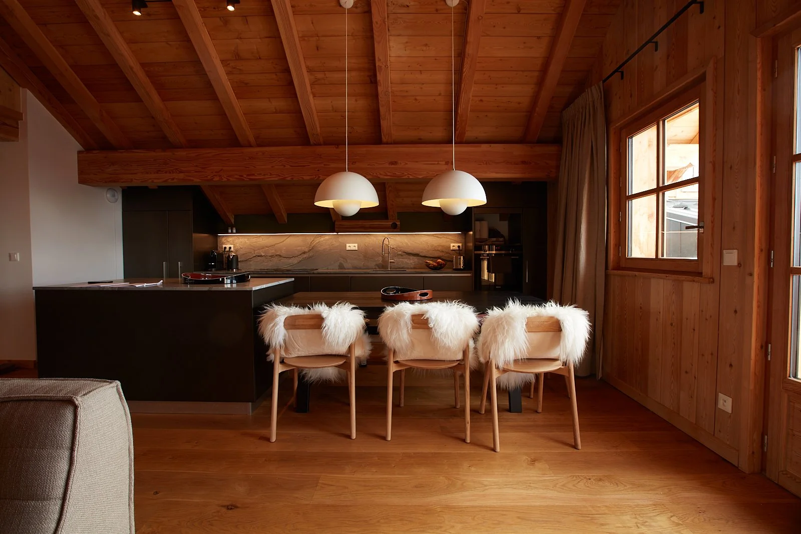 Cuisine en bois avec un grand plan de travail noir, deux lampes suspendues rondes blanches, quatre chaises avec assises en tissu blanc pelucheux, fenêtres en bois laissant entrer la lumière naturelle.