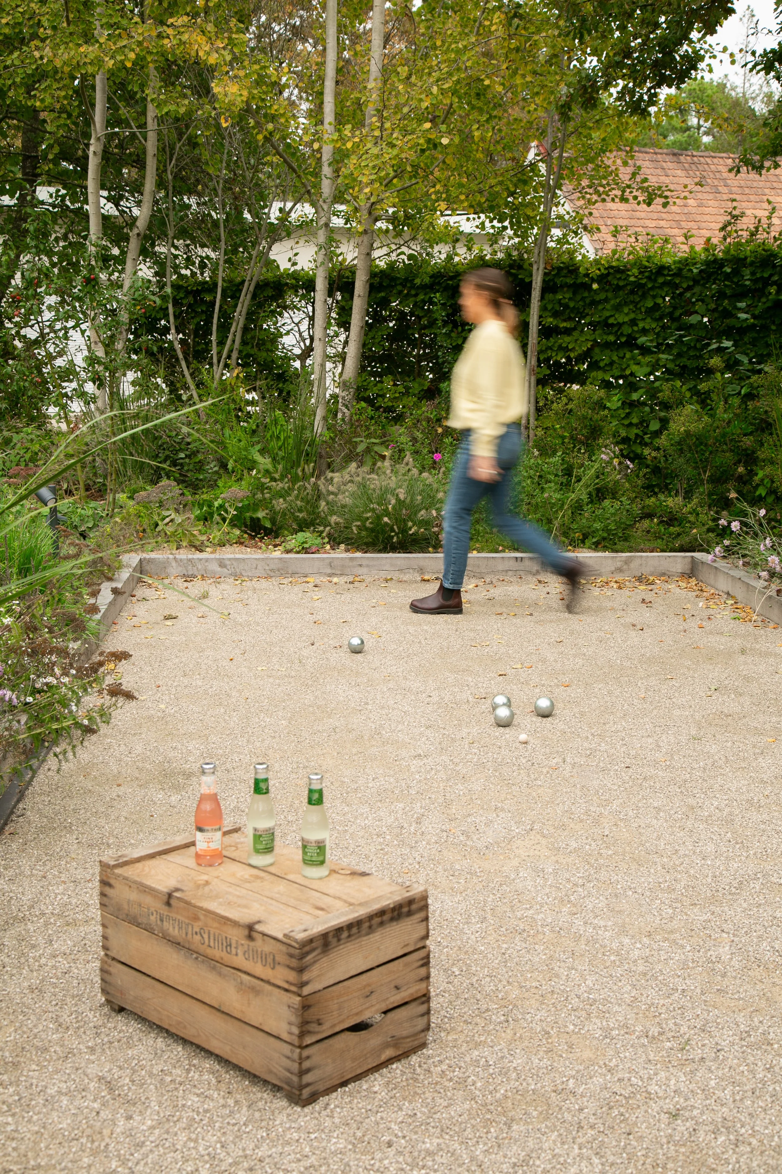 Une femme joue à la pétanque dans un jardin entouré d'arbres et de buissons, avec des bouteilles de boisson posées sur une caisse en bois.