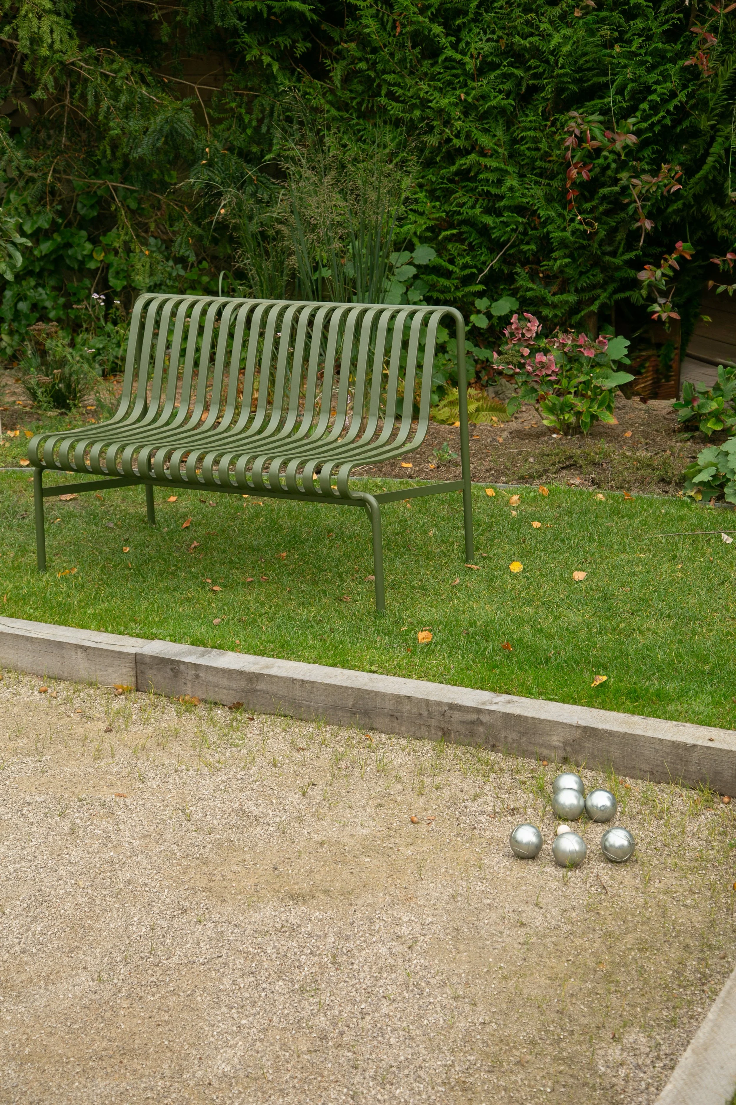 Un banc en métal vert sur une pelouse, à côté d'un terrain de pétanque avec des boules en acier. En arrière-plan, des plantes et des arbustes dans un jardin.
