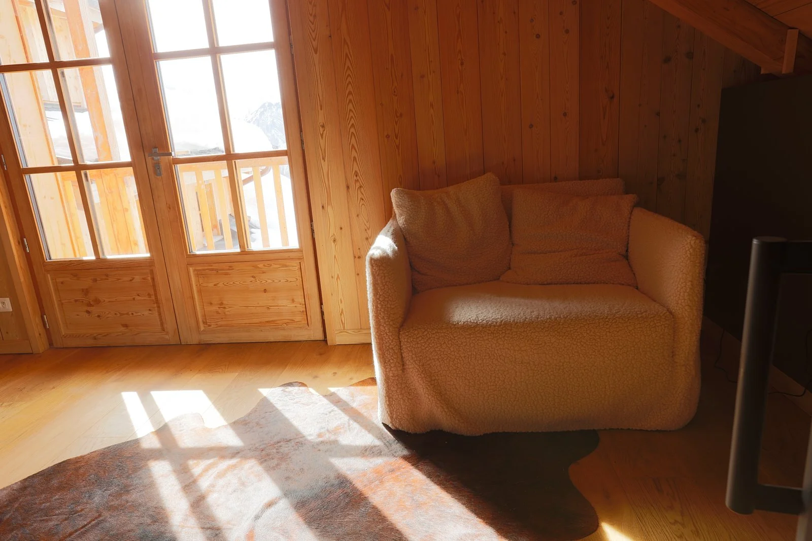 Coin salon avec un canapé beige près d'une porte en bois vitrée, sol en bois, et miroir ou écran noir à droite.