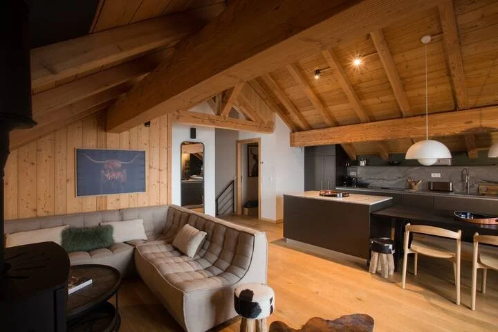 Intérieur d'un salon et cuisine dans une maison avec plafond en bois, canapé beige, cuisine noire moderne, et éclairage suspendu, décor chaleureux.