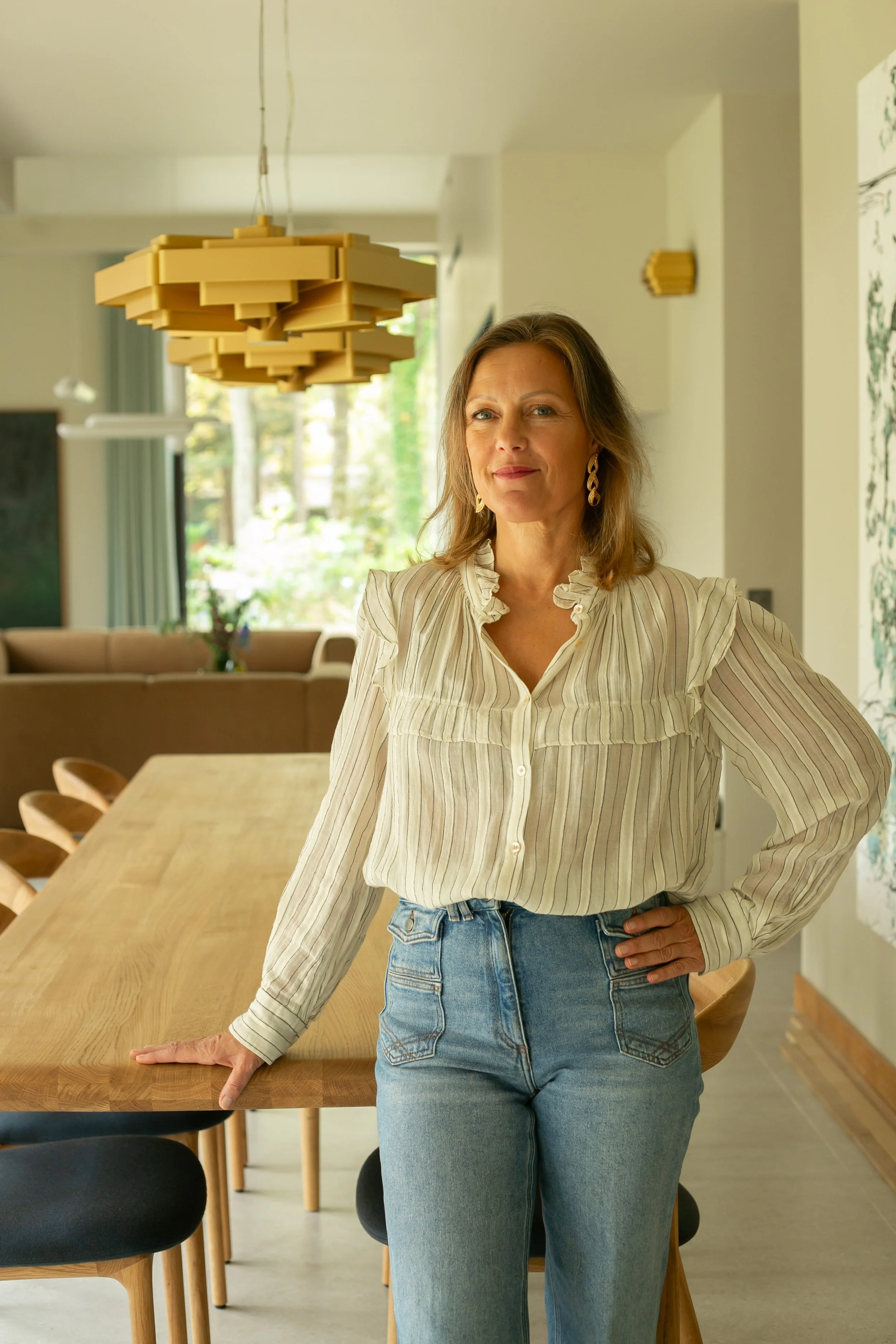 Une femme se tenant près d'une table en bois dans une pièce lumineuse avec beaucoup de lumière naturelle, décorée avec un éclairage suspendu en bois et des œuvres d'art sur le mur.