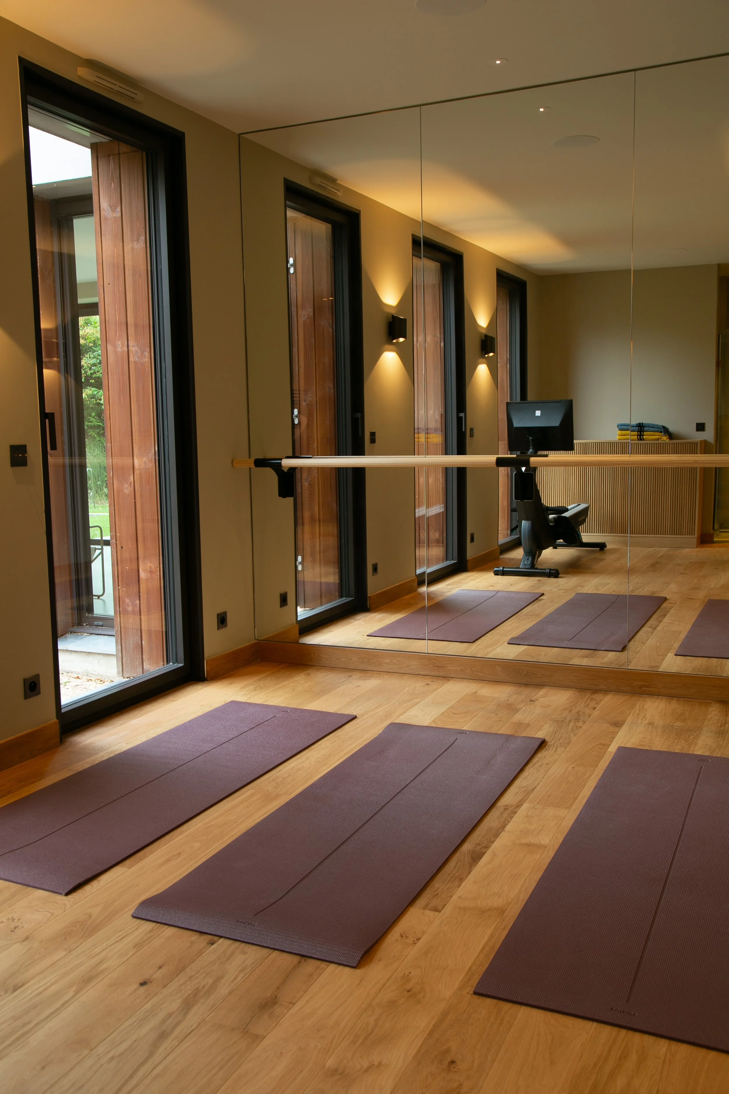 Salle de yoga avec tapis marron, une barre de ballet fixée au mur miroir, un bureau avec un ordinateur et un fauteuil, éclairage tamisé et grande fenêtre laissant entrer la lumière naturelle.