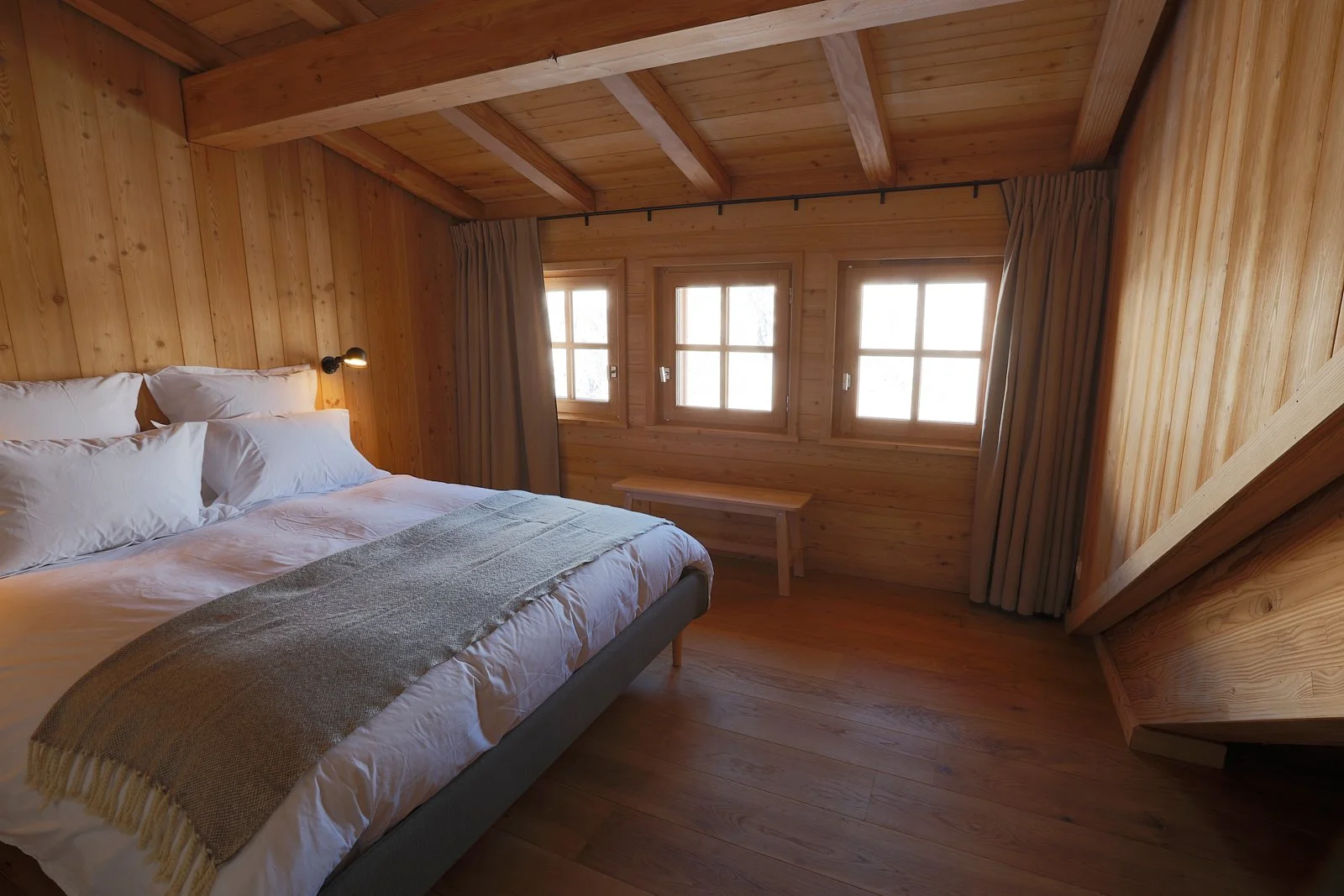 Chambre en bois avec un lit fait, trois fenêtres, un banc en bois sous les fenêtres, et des rideaux beige.