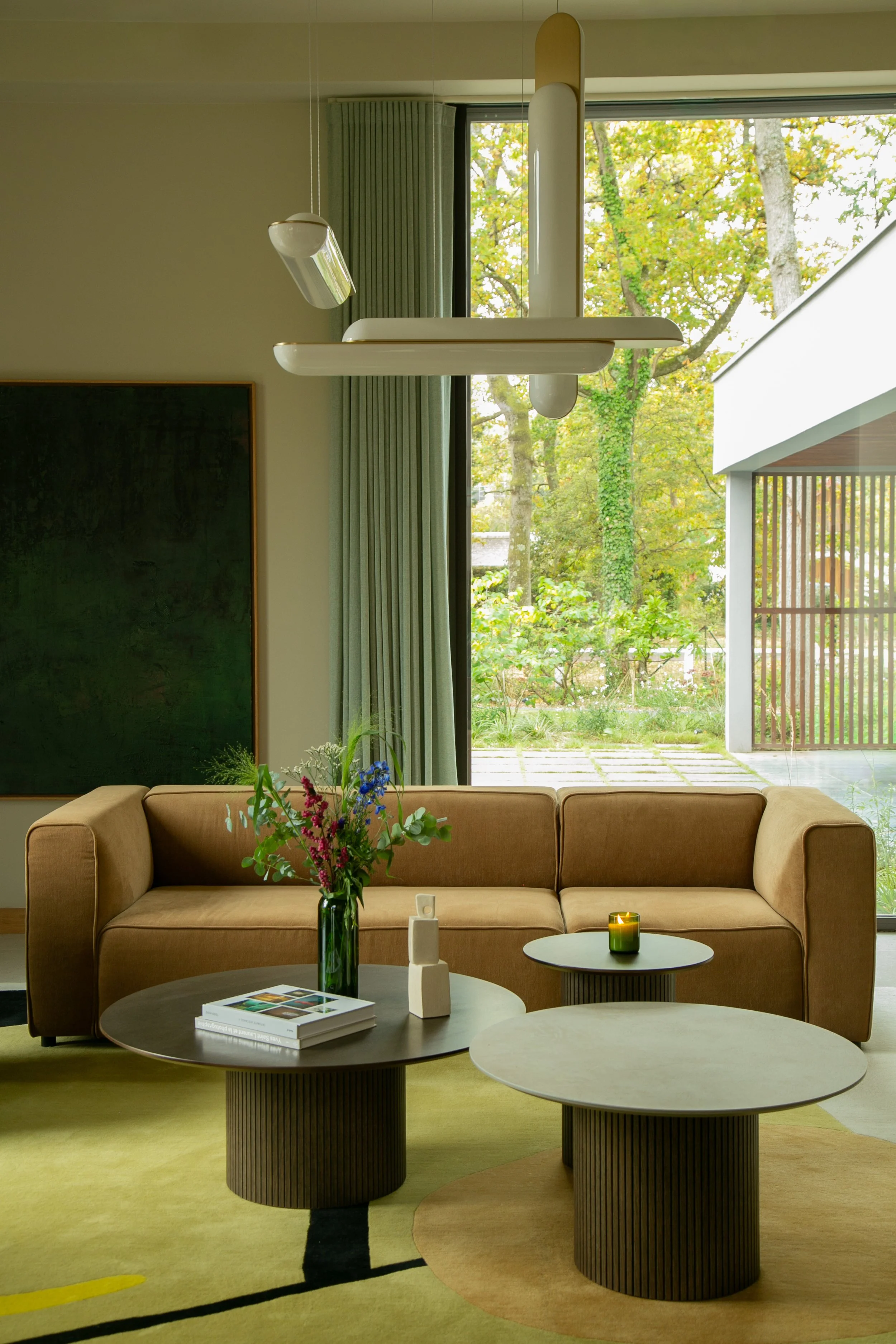 Salon moderne avec un canapé beige, tables basses rondes en bois, vase avec des fleurs, bougie verte, devant une grande fenêtre avec vue sur un jardin avec des arbres.