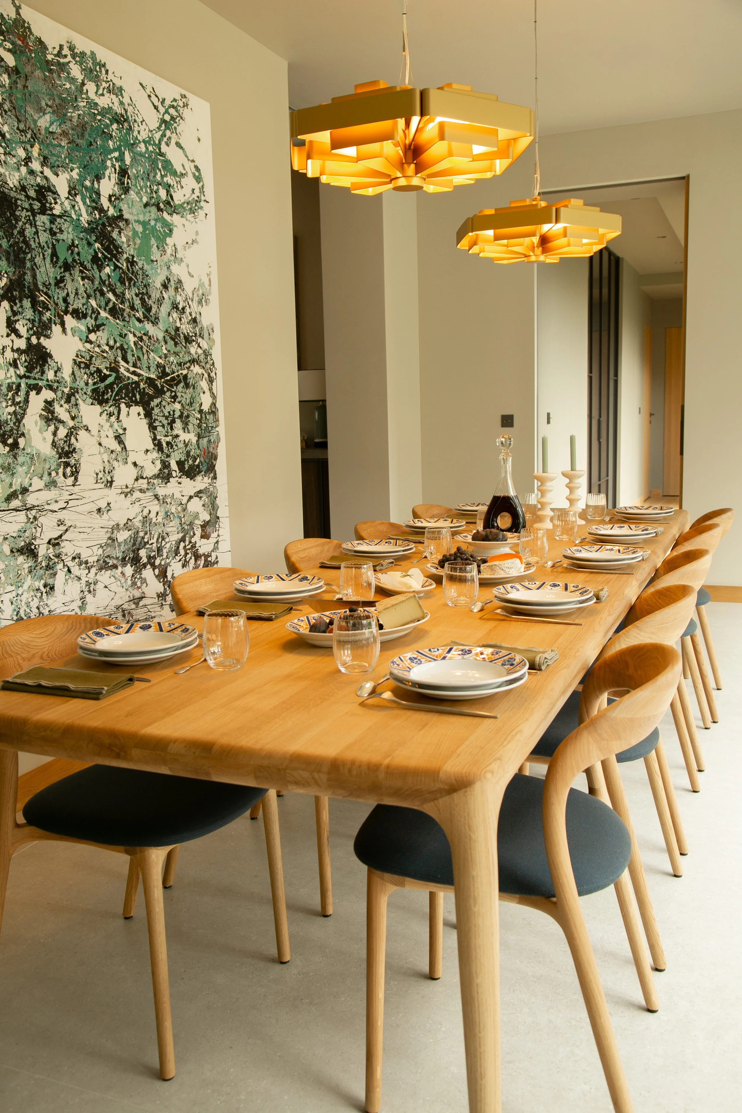 Une salle à manger moderne avec une grande table en bois entourée de chaises en bois avec coussins noirs, décorée pour un repas avec des assiettes, verres, couverts, bougies et un drapeau en verre au centre, sous deux luminaires suspendus en forme de