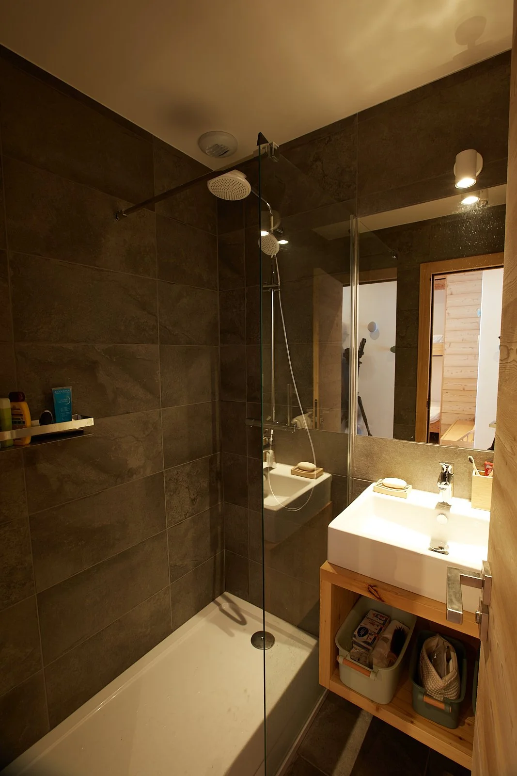 Salle de bain avec douche à l'italienne, lavabo blanc, miroir, accessoires de toilette et éléments en bois, murs en tuiles foncées et éclairage modéré.