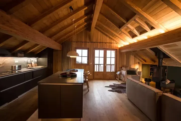 Intérieur d'une pièce à l'allure chaleureuse en bois, avec une cuisine moderne à gauche, un coin salon avec canapé à droite, et de grandes fenêtres en arrière apportant de la lumière naturelle.