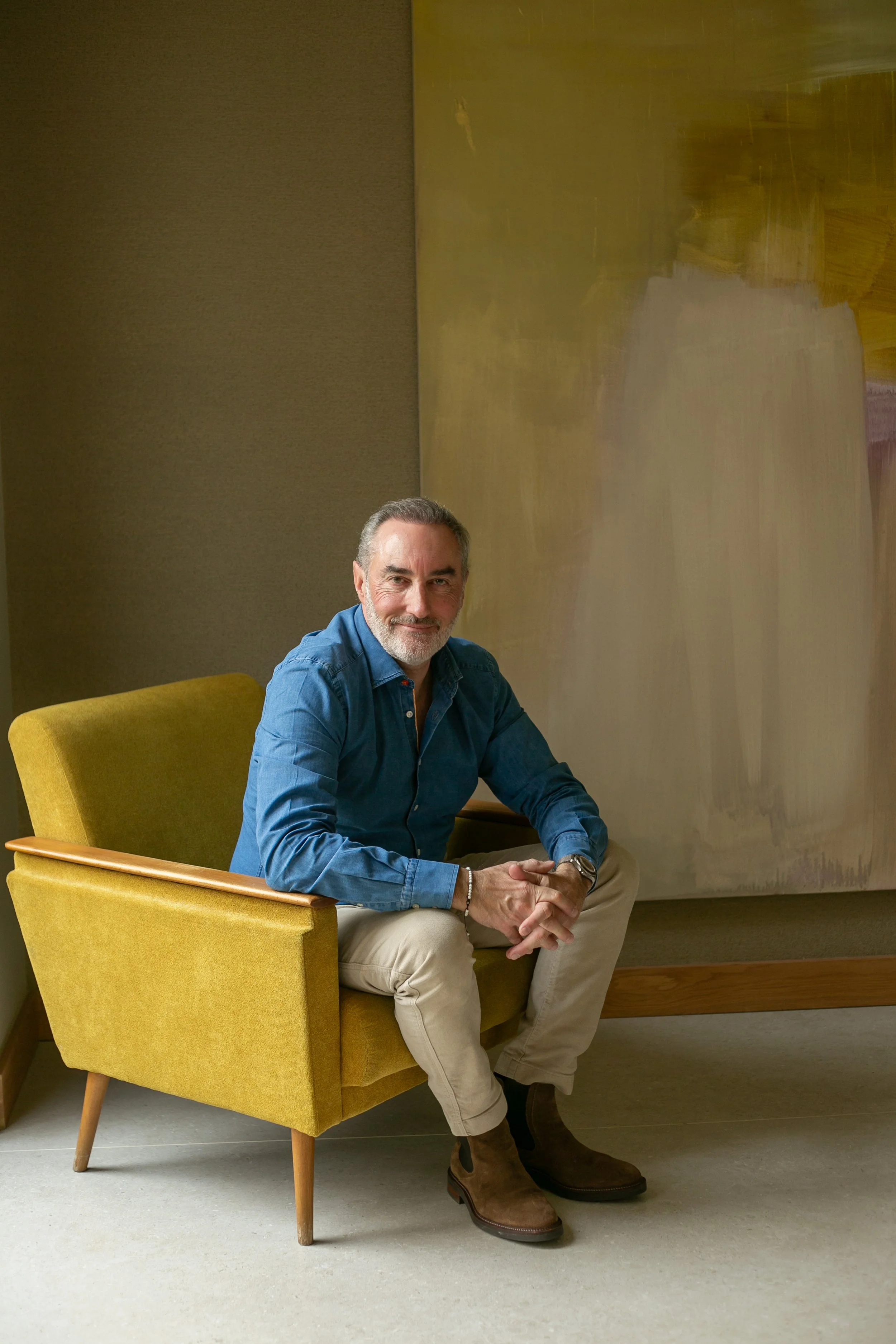 Un homme souriant assis dans un fauteuil jaune, portant une chemise bleue et des bottines marron, dans une pièce avec un mur en peinture abstraite.