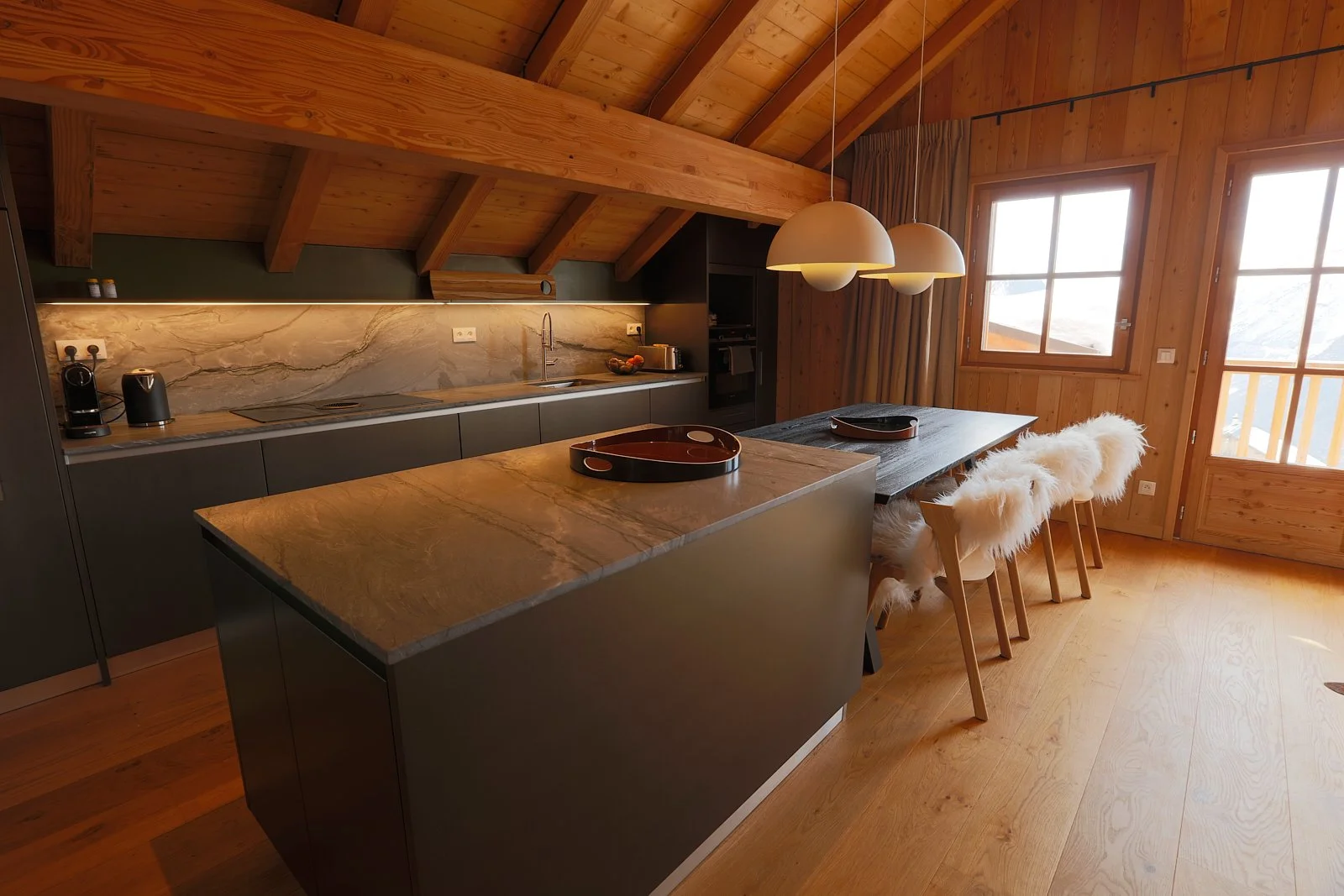 Cuisine moderne en bois avec îlot central, deux suspensions lumineuses, table à manger en bois avec quatre chaises avec coussins en fausse fourrure, fenêtres laissant entrer la lumière naturelle, décor rustique-chic.