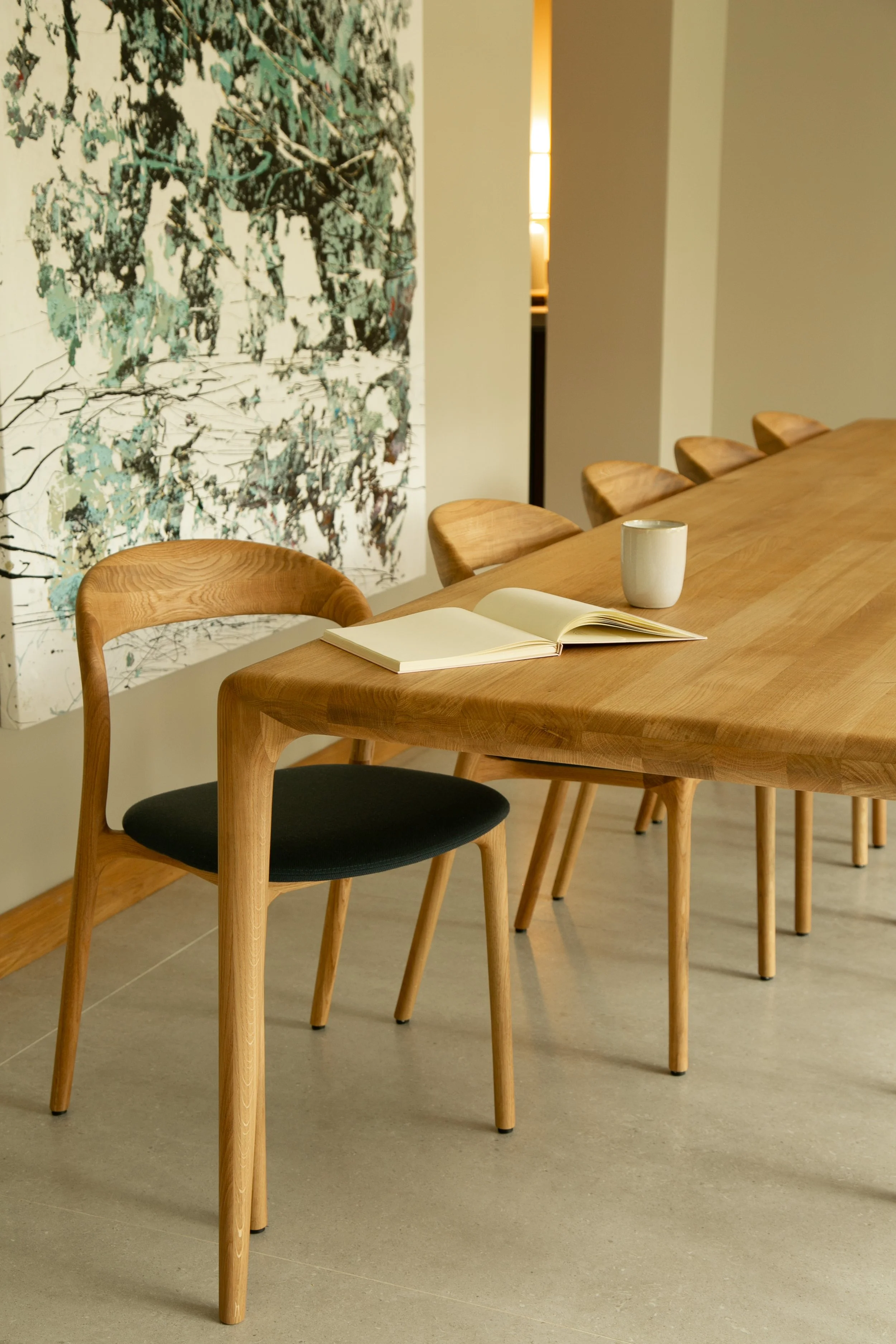 Une pièce avec une grande table en bois, entourée de chaises en bois, un livre ouvert et une tasse blanche sur la table, décorée d'une œuvre d'art abstraite au mur et de murs de couleur claire.