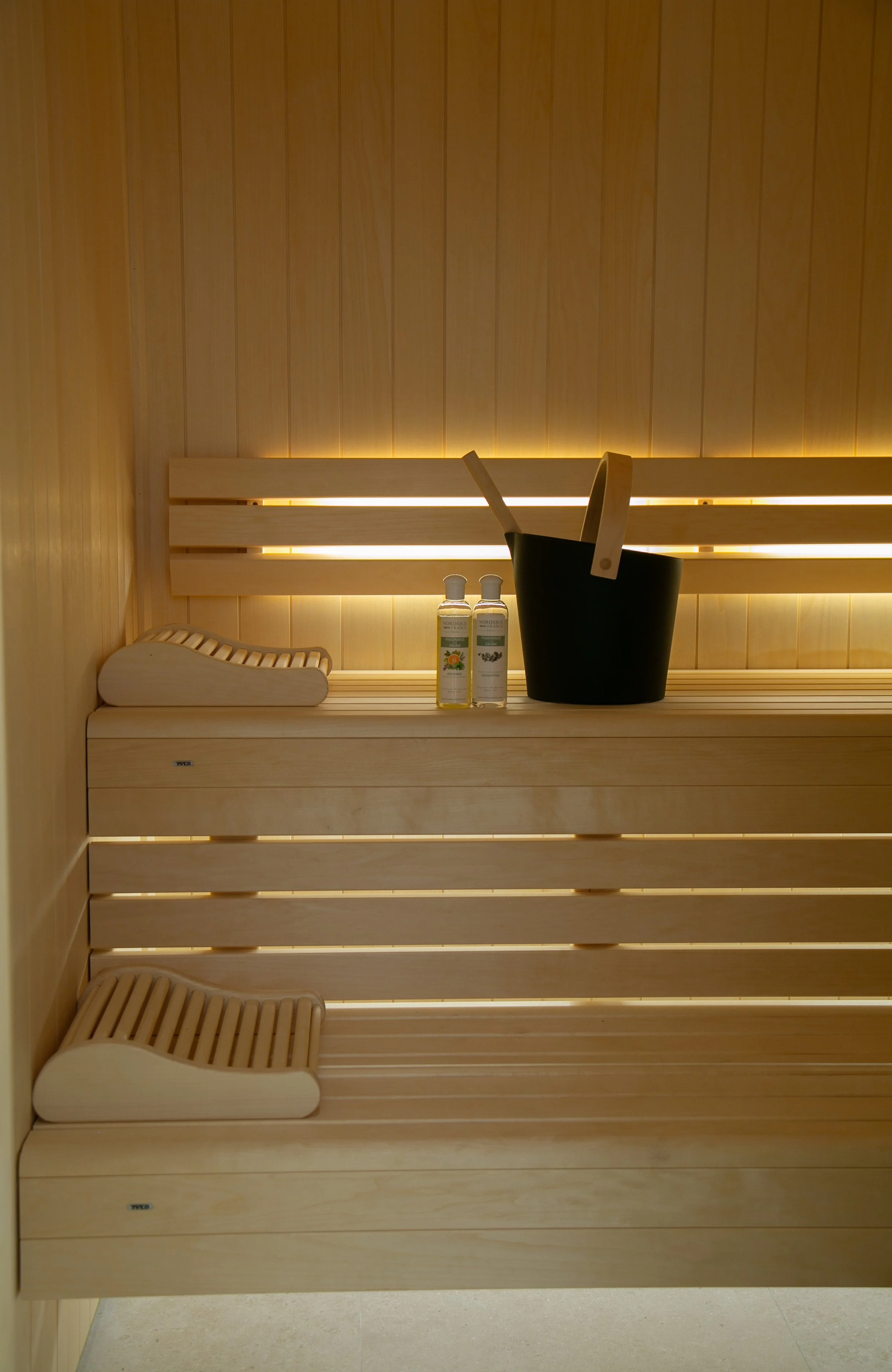 Une cabine de sauna en bois avec deux blocs de bancs en haut et en bas, deux bouteilles d'huile ou d'aromathérapie sur le banc du haut, un seau en bois avec deux manches pour la chaudière ou le bain de vapeur.