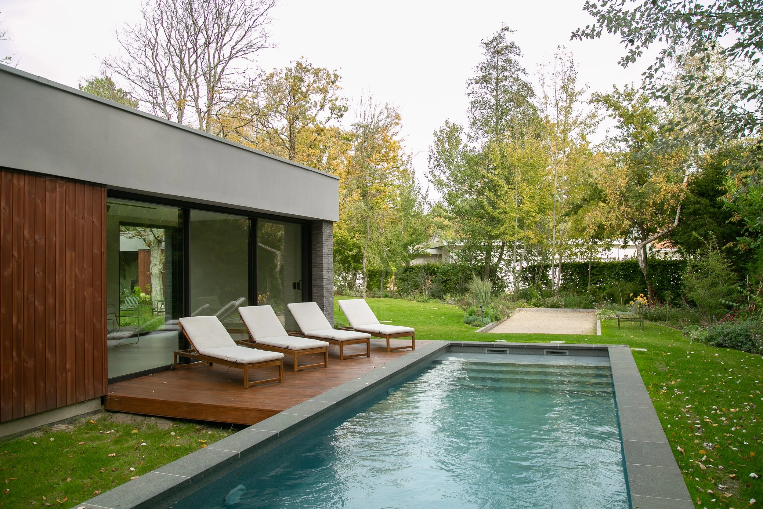 Une maison moderne avec une piscine, un patio en bois, quatre chaises longues blanches, et un jardin arboré.