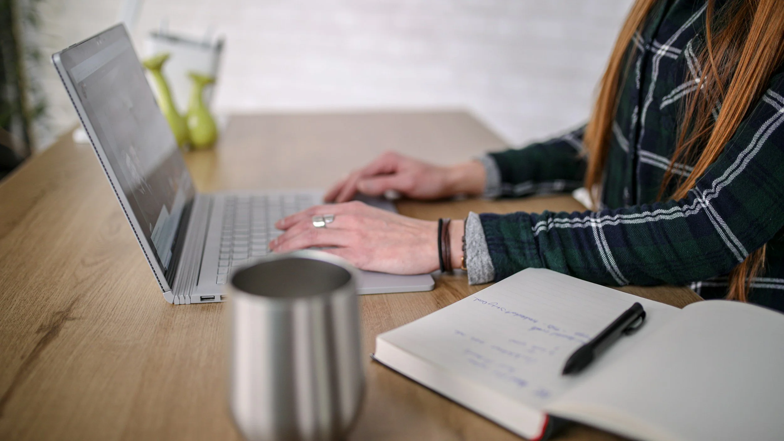Person with long red hair working on a laptop at a wooden desk, with a notebook and pen nearby.