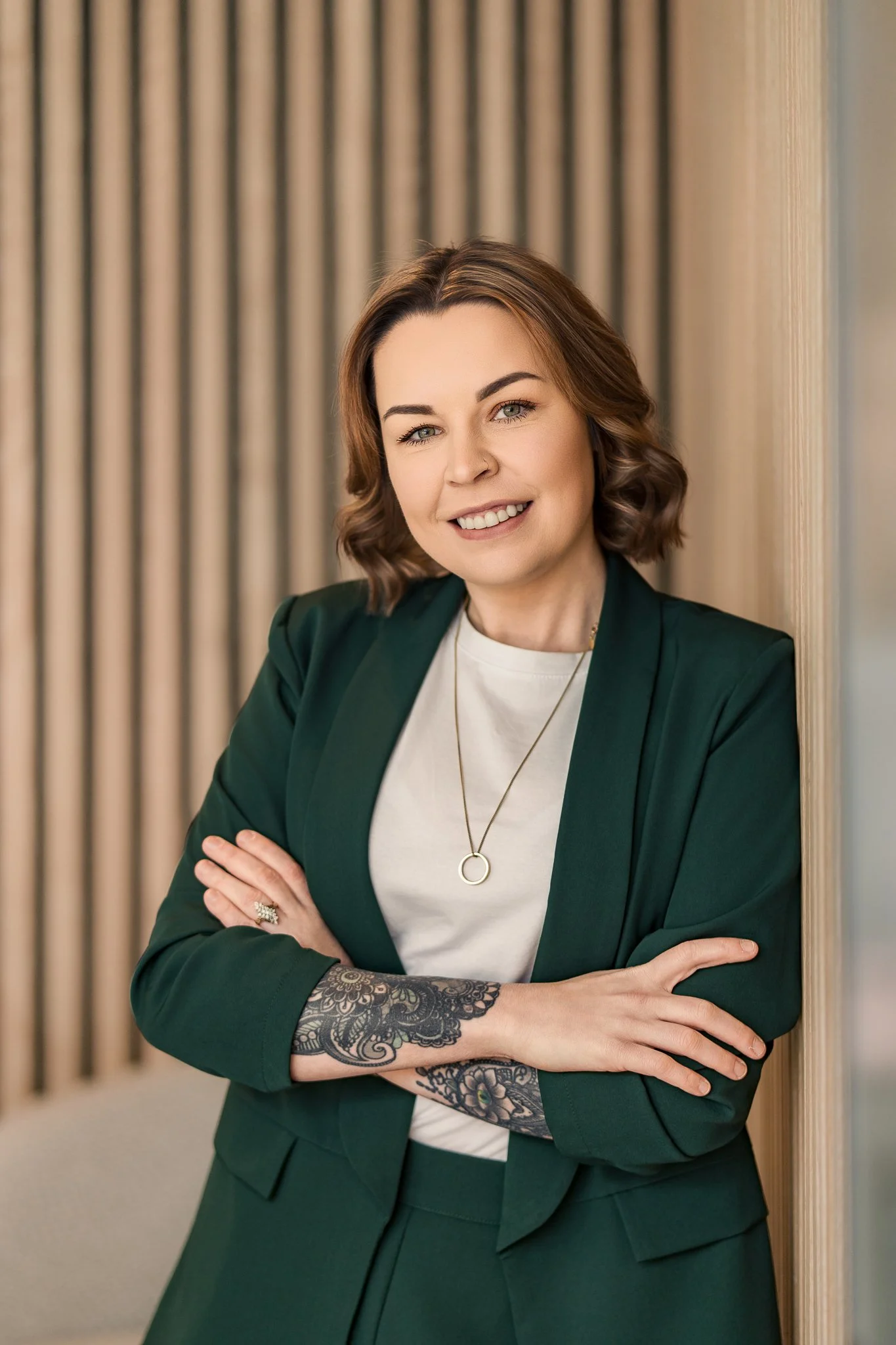A woman with short, wavy brown hair and light skin leaning against a wall, smiling, wearing a dark green blazer over a white top, with a tattoo on her left forearm, and a necklace with a circular pendant.