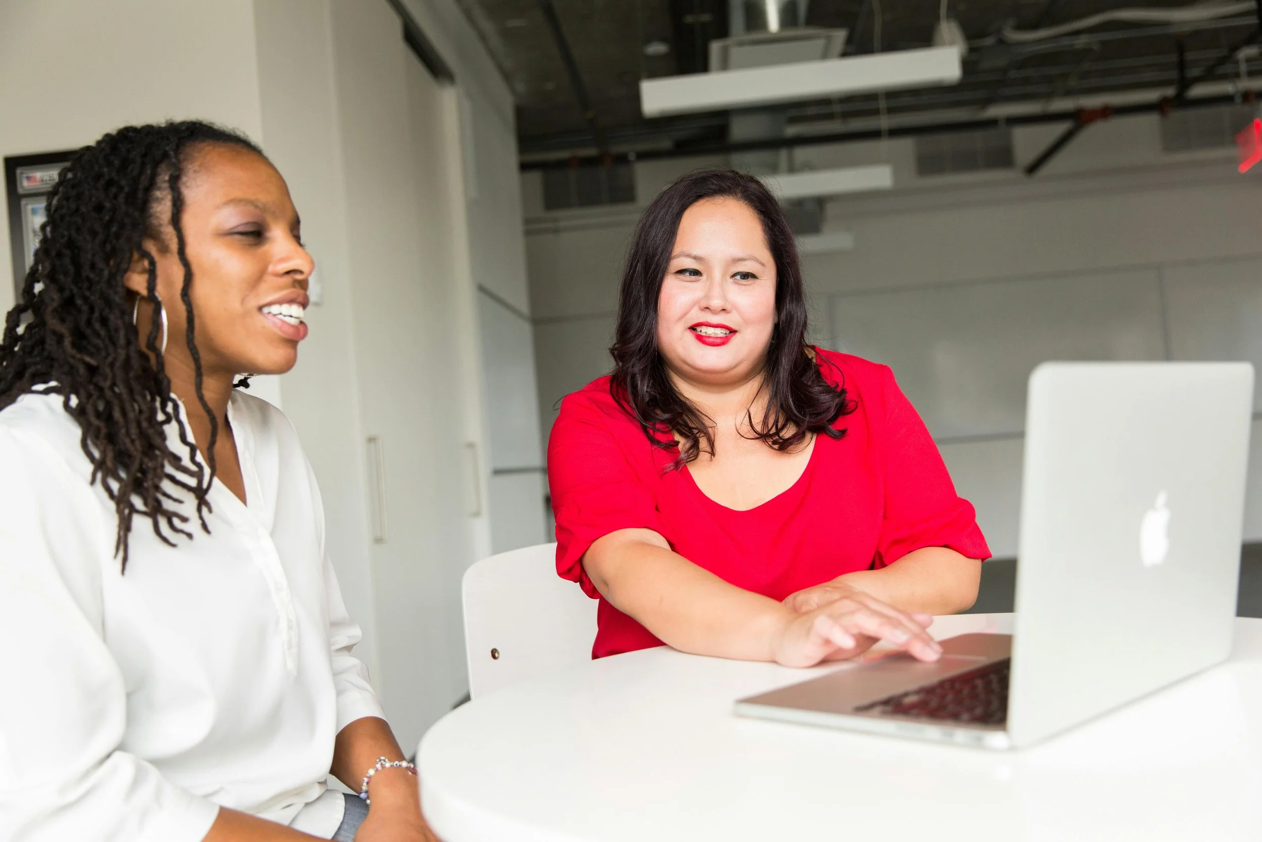 Two women sitting at a white table with a laptop, having a discussion in a modern office environment.