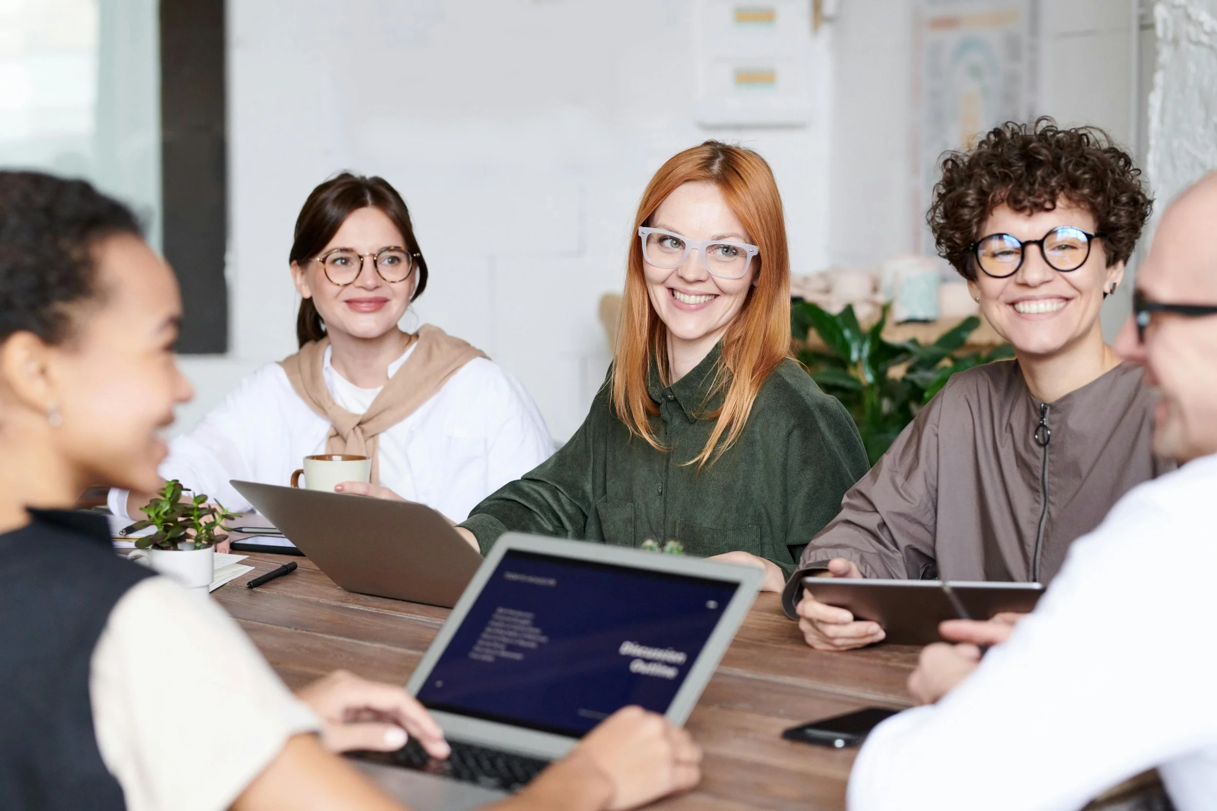 Group of five diverse women sitting around a wooden table in a modern office, smiling and engaging in a meeting, with laptops, tablets, notebooks, cups, and a small potted plant on the table.