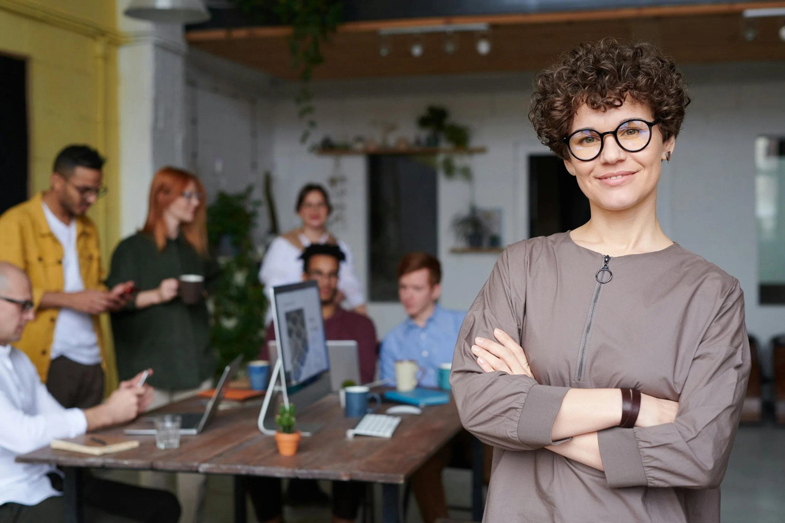 A woman with short curly hair, glasses, and a gray top smiling with arms crossed in a modern office setting. In the background, a diverse group of people are working at a table with laptops and coffee cups.