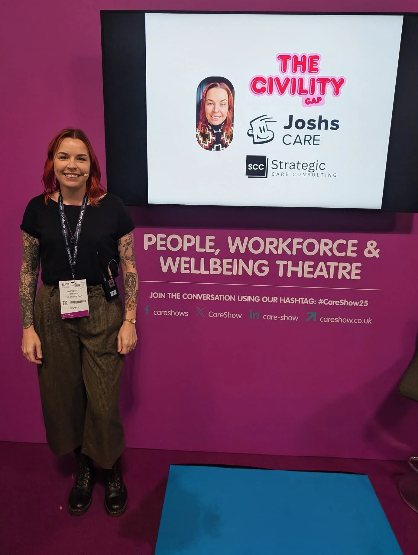 A woman with shoulder-length red hair and tattoos on her arms is standing in front of a purple wall with a large screen displaying a presentation slide. The slide shows a cartoon image of a woman, the text "The Civility Gap," and logos for Joshs Care and Strategic Care Consulting.