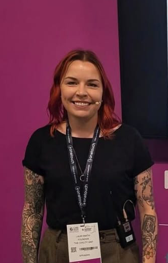 Smiling woman with reddish hair, tattoos on arms, wearing a black shirt, conference badge, and a headset microphone, standing against purple and dark blue background.