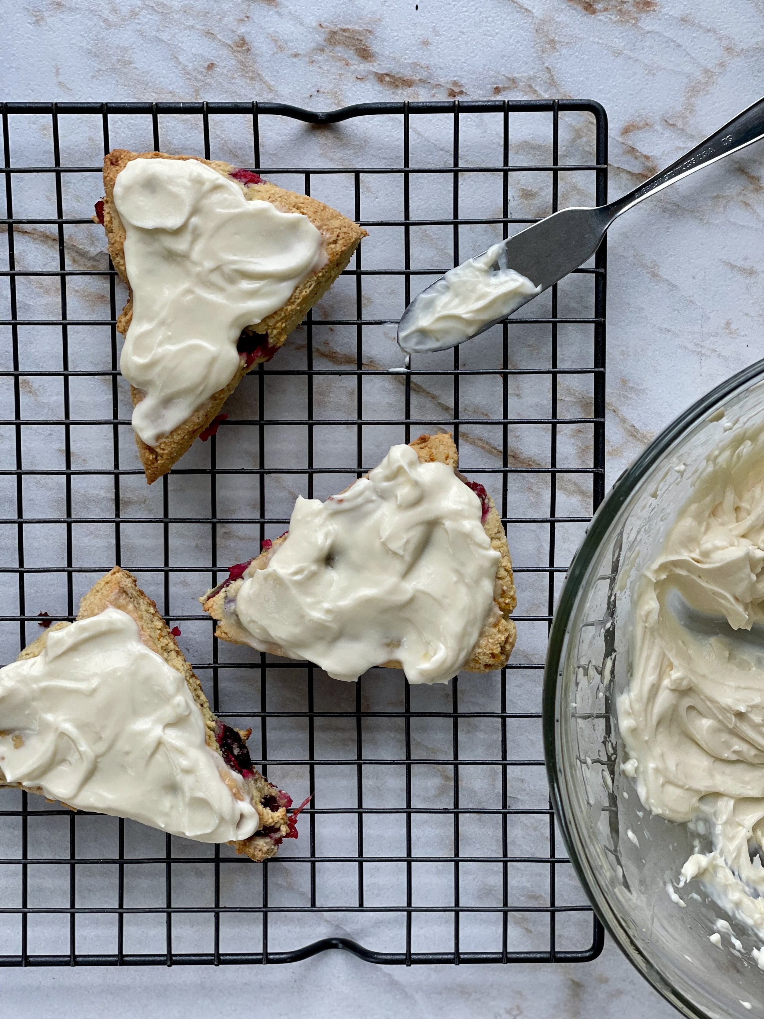 Slices of a berry crumb cake with white frosting on a cooling rack, with a bowl of frosting nearby and a spatula with frosting on it.