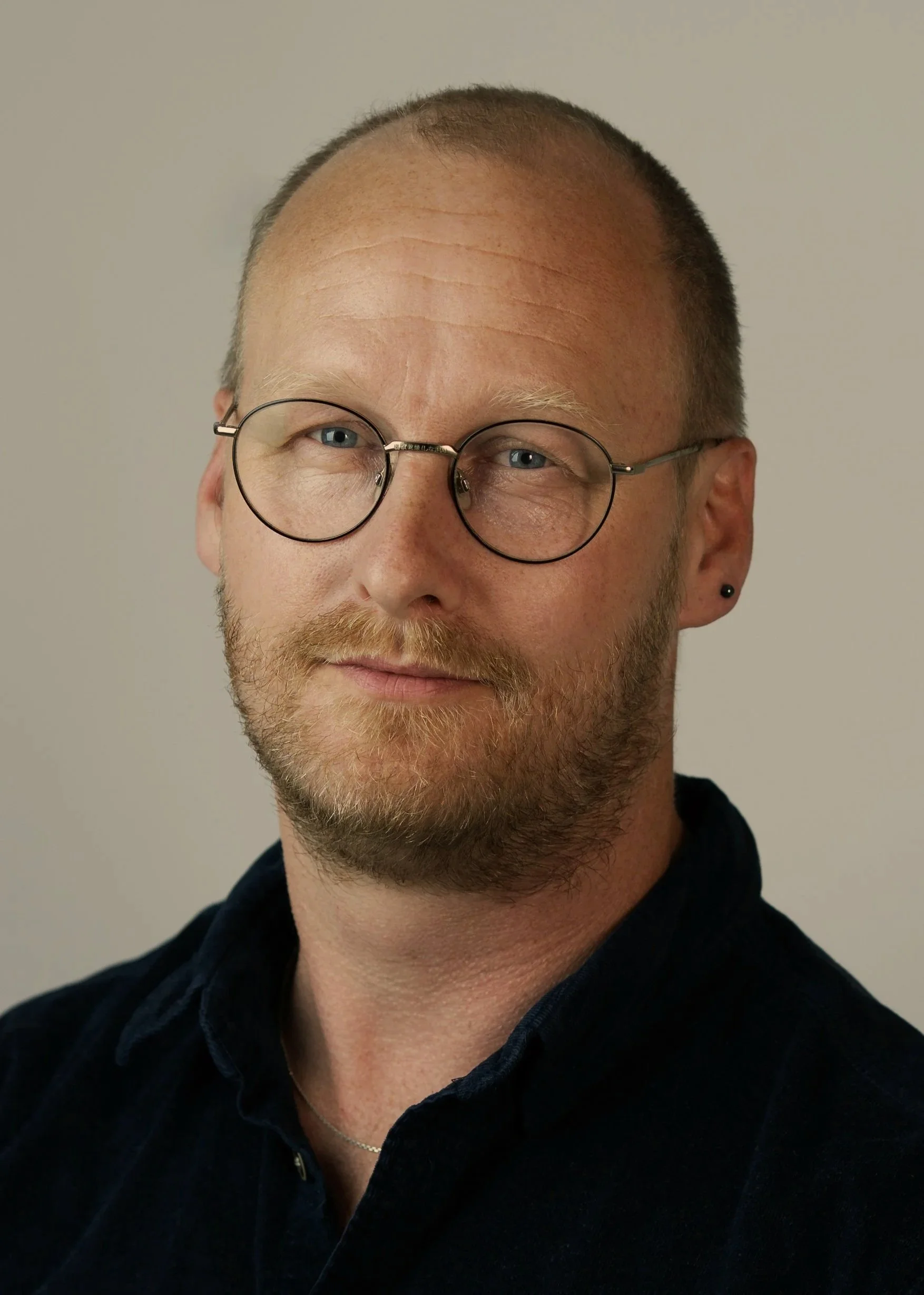 A close-up portrait of a man with a shaved head and beard, wearing black-rimmed glasses and a black shirt, smiling softly against a plain background.