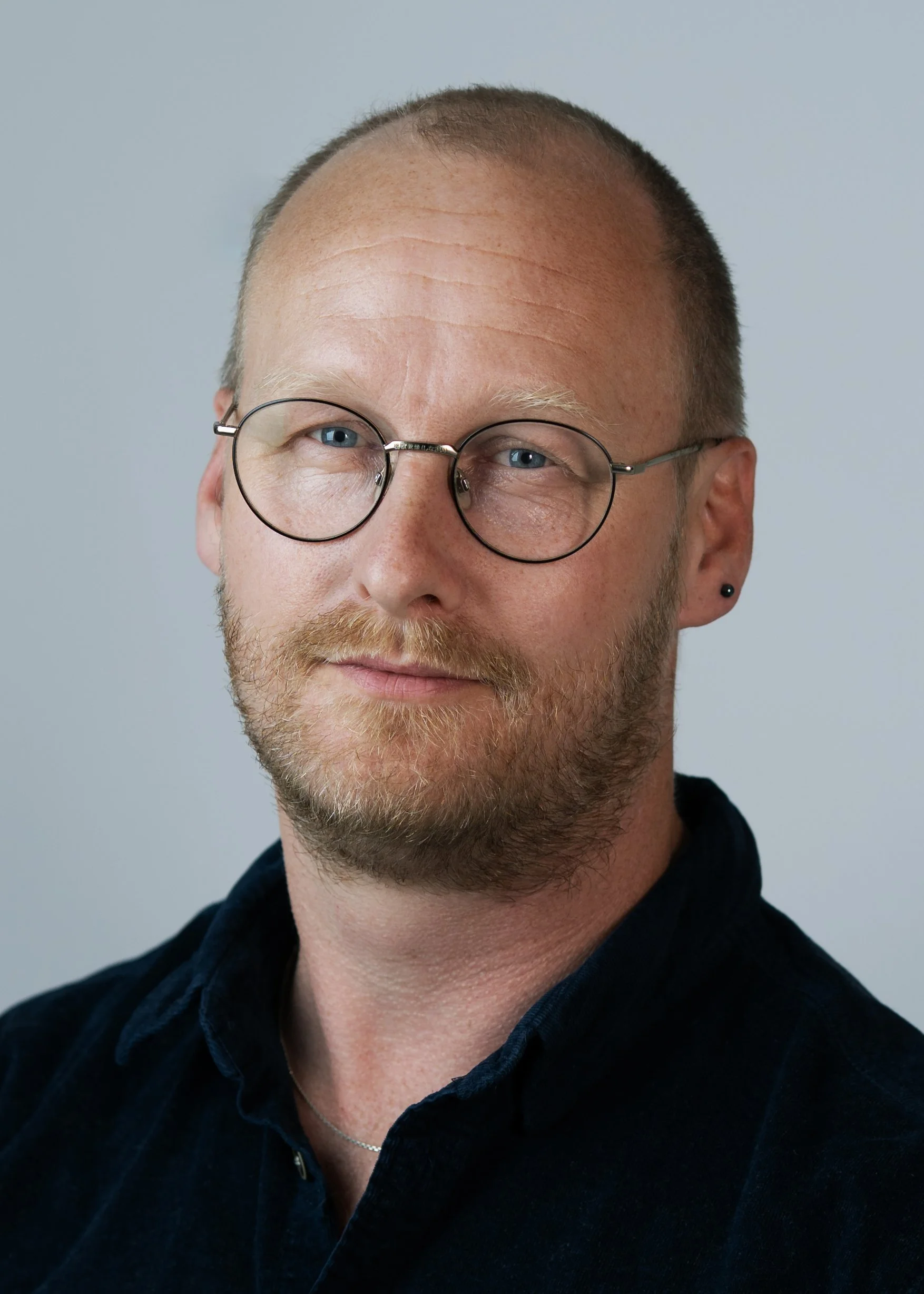 A close-up of a man with glasses and a beard, smiling, wearing a black shirt, against a plain background.