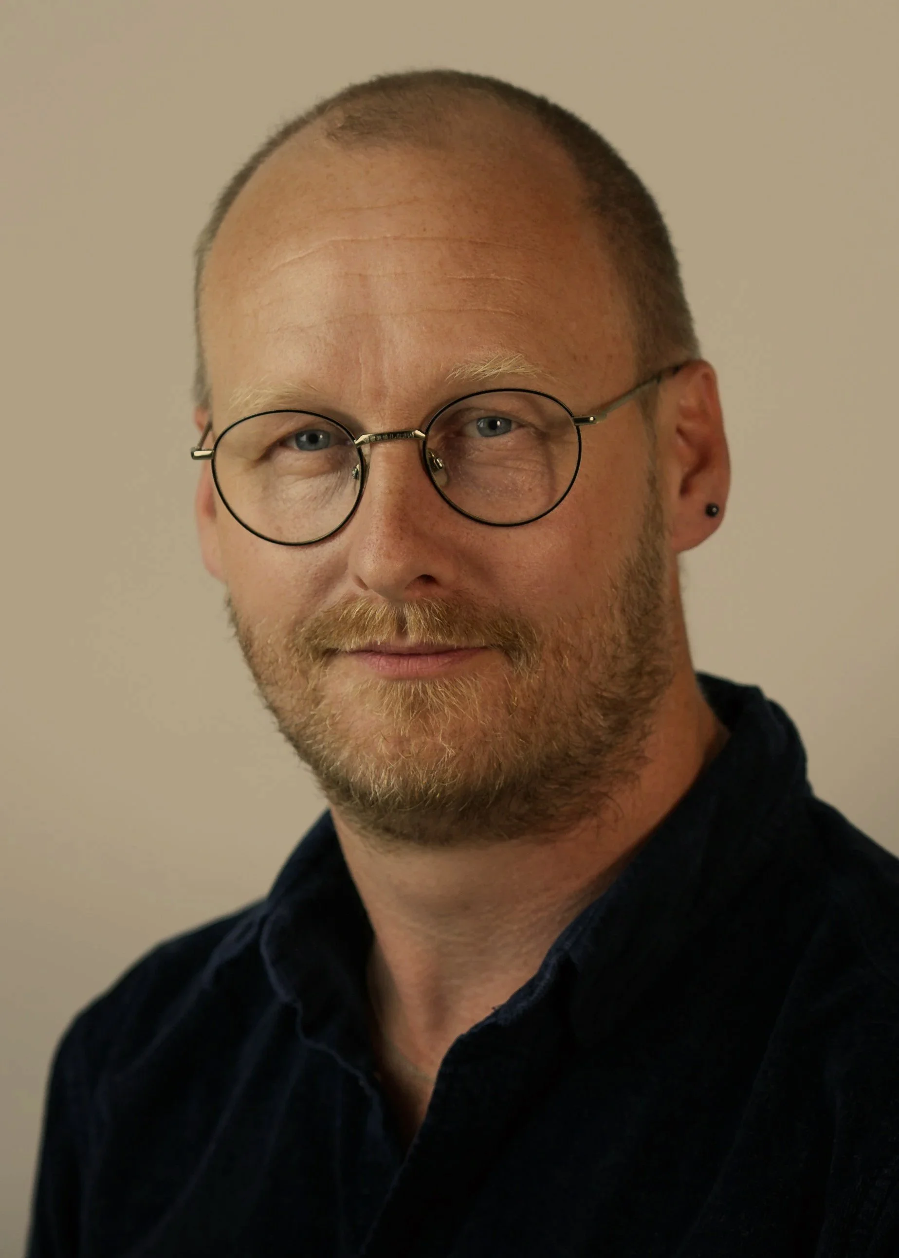Close-up of a man with light skin, short beard, wearing round glasses and a dark shirt, standing against a plain background.