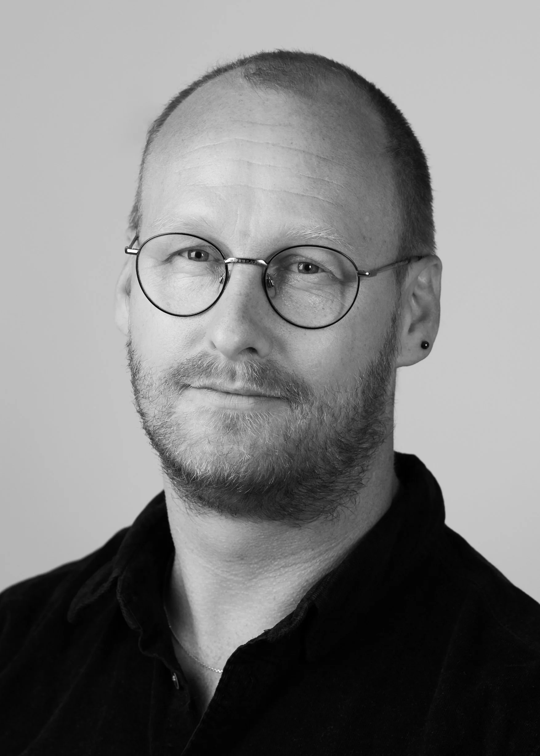 Black and white portrait of a man with glasses, short hair, beard, and wearing a dark shirt and small earring.