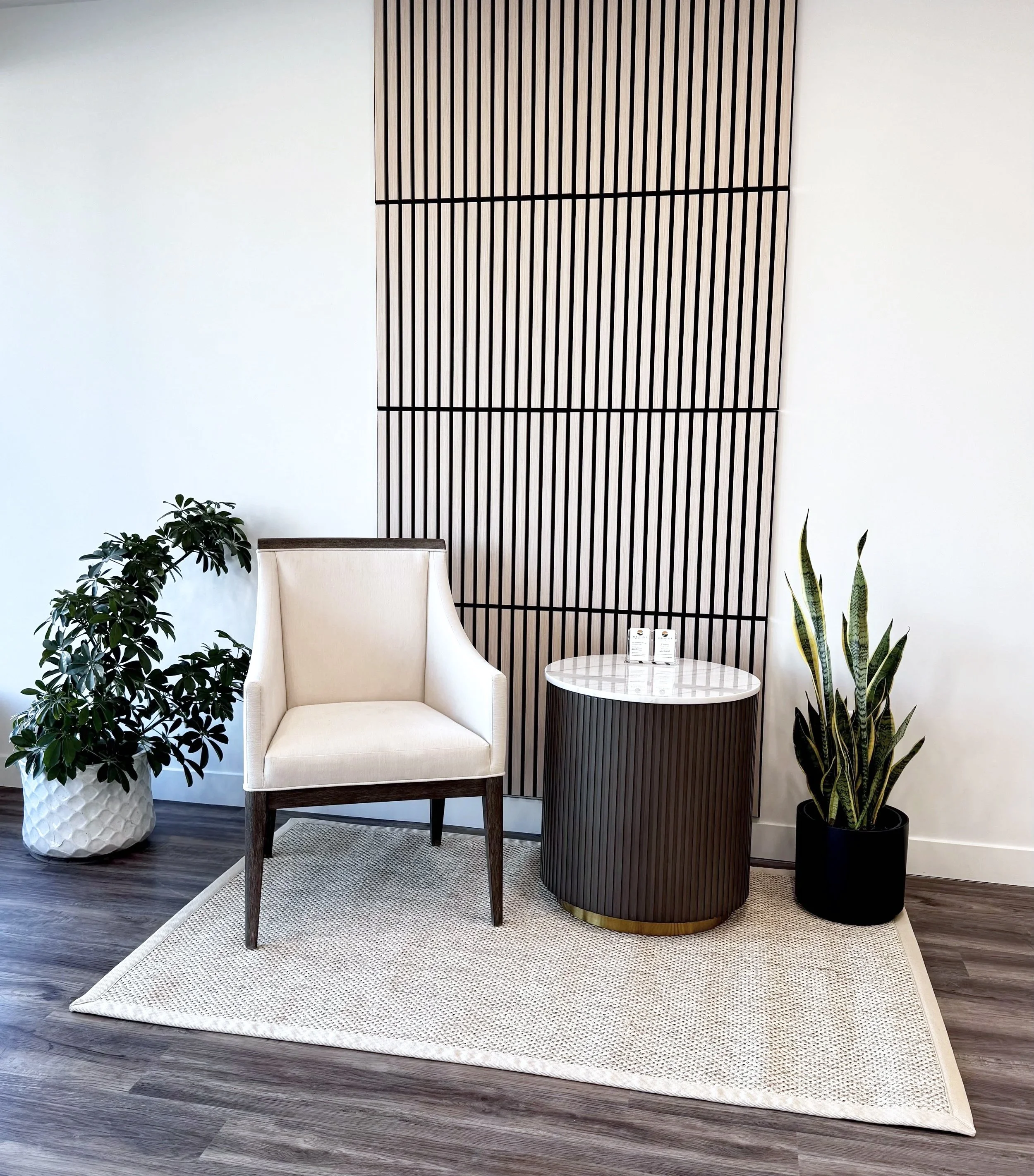 Modern memorial showroom seating area with white armchair, small round table, potted plants, and a decorative wooden wall panel.
