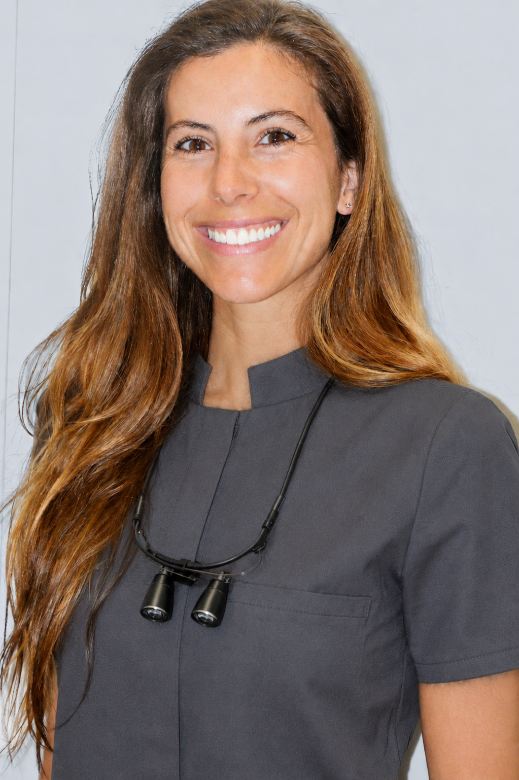 Médica o dentista con bata gris y lupa en el cuello, sonriendo frente a fondo blanco.