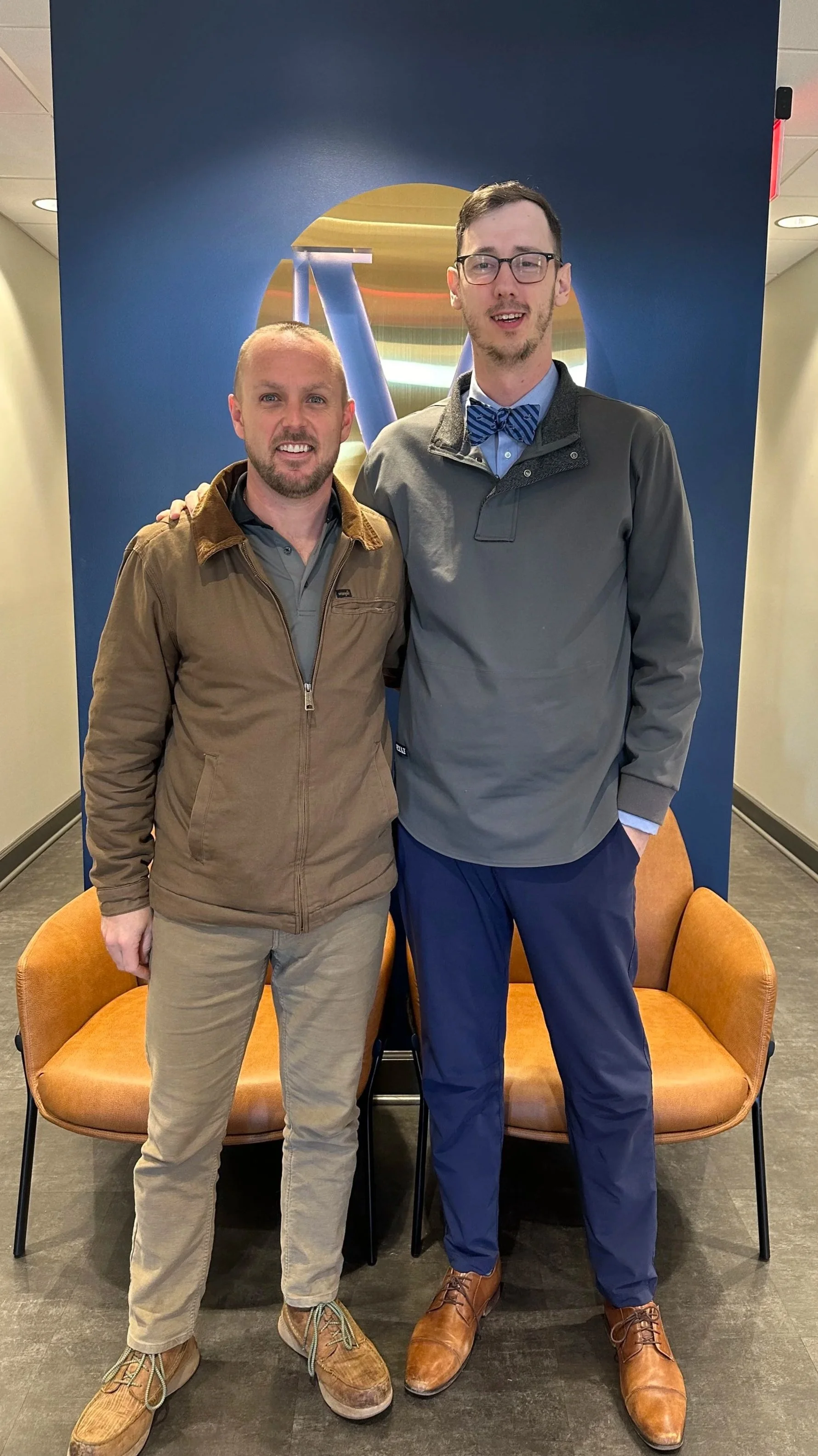 Two men standing on a bench in front of a blue wall with a colorful abstract logo. One man has a beard, is wearing a tan jacket, and khaki pants. The other man is taller, wearing glasses, a bow tie, a gray jacket, and navy blue pants. They are smiling and have their arms around each other.