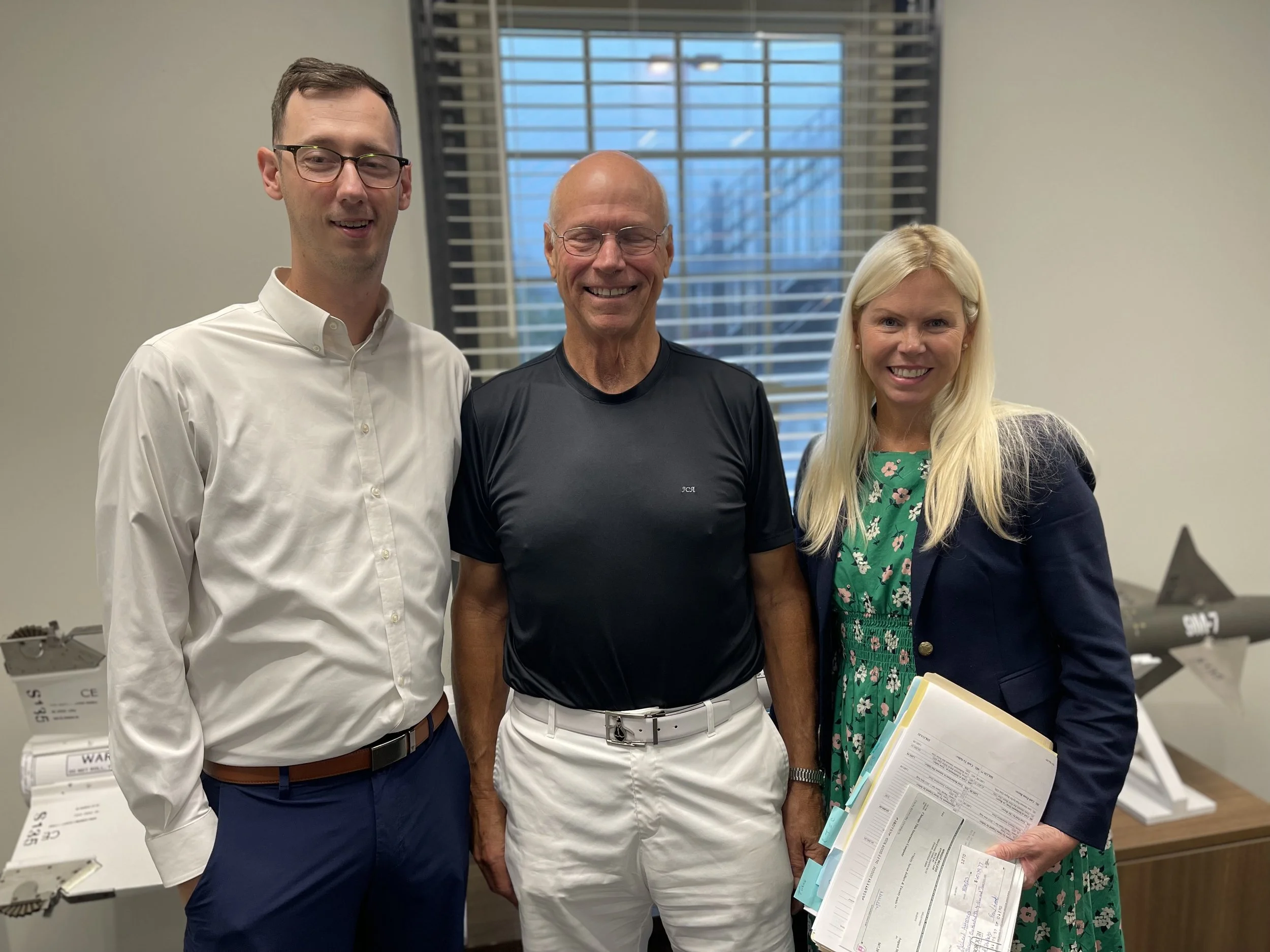 Three people standing together indoors, smiling, with documents and office equipment in the background.