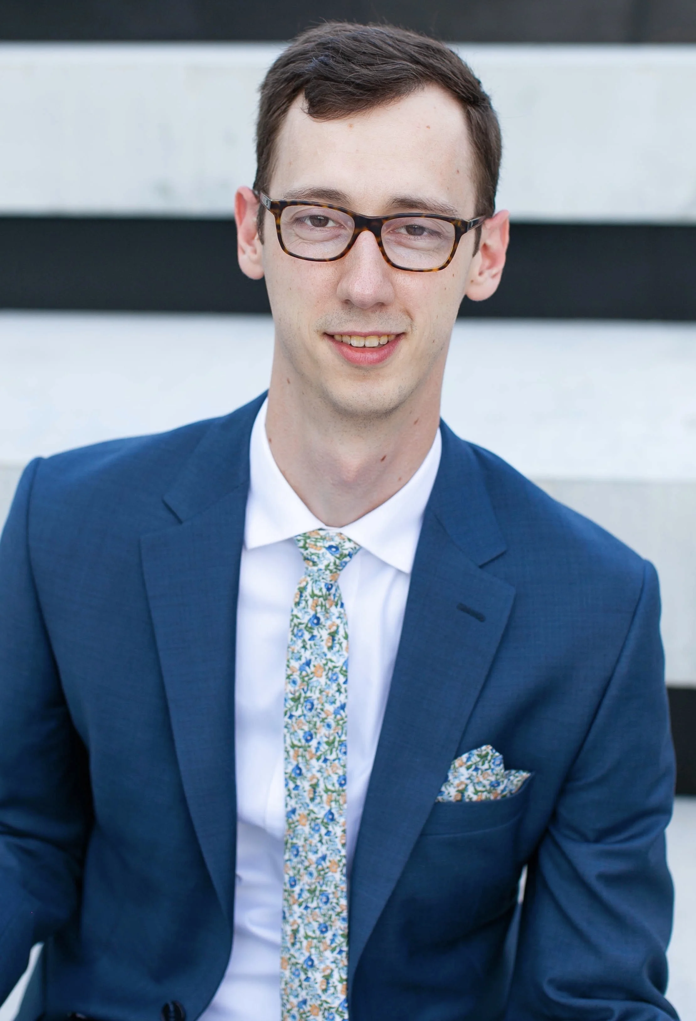 A young man with short brown hair, wearing glasses, a navy blue suit, a white shirt, and a floral tie and pocket square, sitting outside against a grey and black striped background.