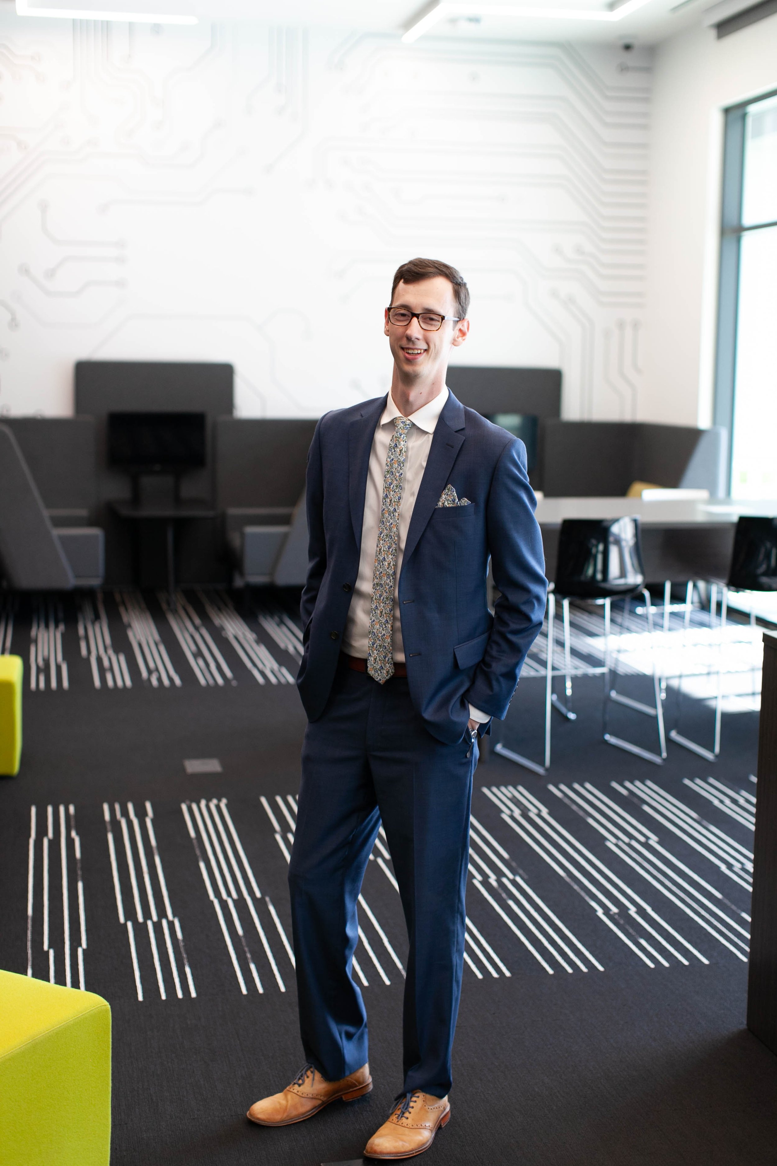 A man in a business suit with glasses standing in a modern office, smiling with hands in pockets, with black office chairs and a wall with a circuit board pattern in the background.