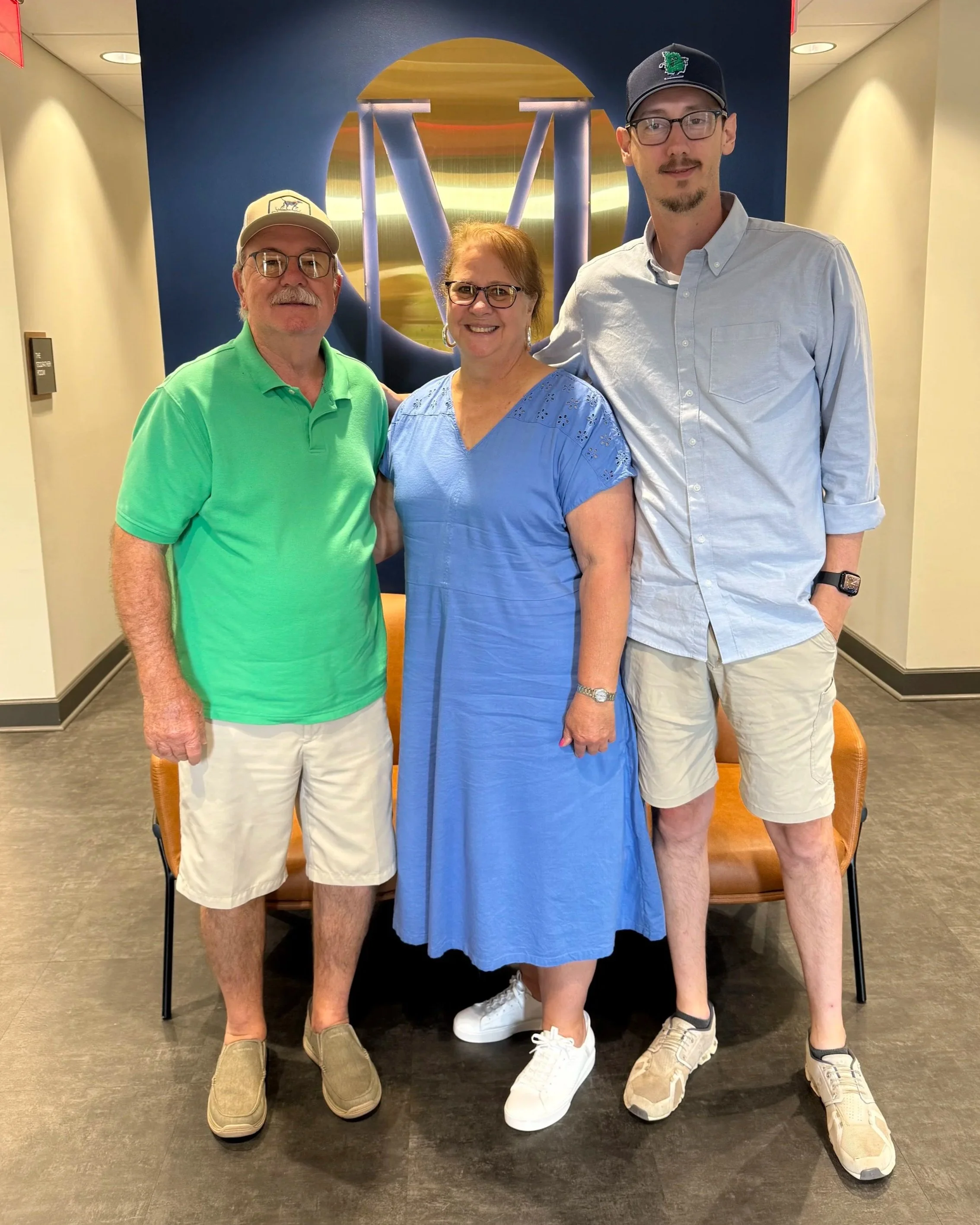 Three people standing together in front of a large art piece with a metallic globe and blue background. The group includes two men and one woman, all wearing casual clothing and glasses.
