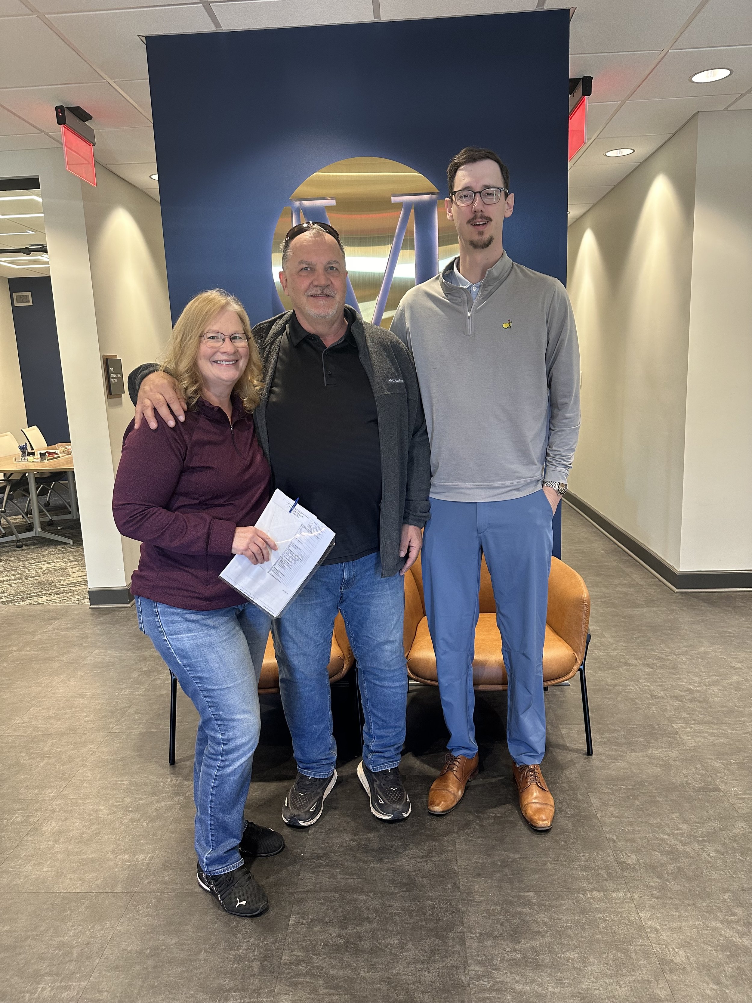 Three people standing together indoors, posing for a photo in front of a blue wall with a logo. The person on the left is a woman wearing glasses, a maroon jacket, and jeans, holding papers. The middle person is a man with glasses, a black shirt, gray jacket, and jeans. The person on the right is a tall man with glasses, a gray quarter-zip pullover, light blue pants, and brown shoes.