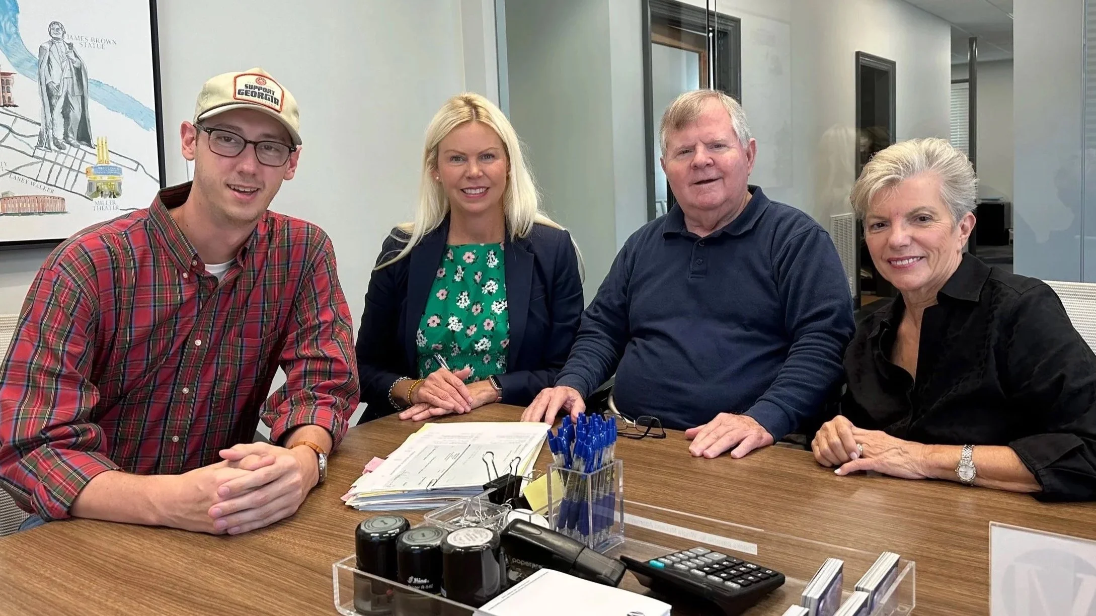 Four people sitting at a conference table with papers, pens, and office supplies, smiling for a photo in a well-lit office.