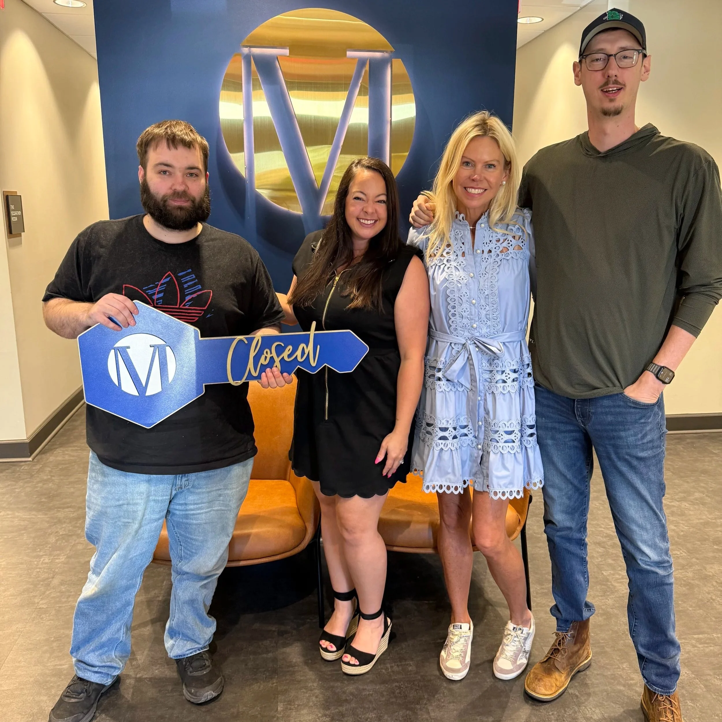 A group of five people, four men and one woman, standing close together inside a building with a blue and gold logo behind them that says 'Closed'. The group is smiling, and the woman is holding a sign with the word 'Closed' on it.