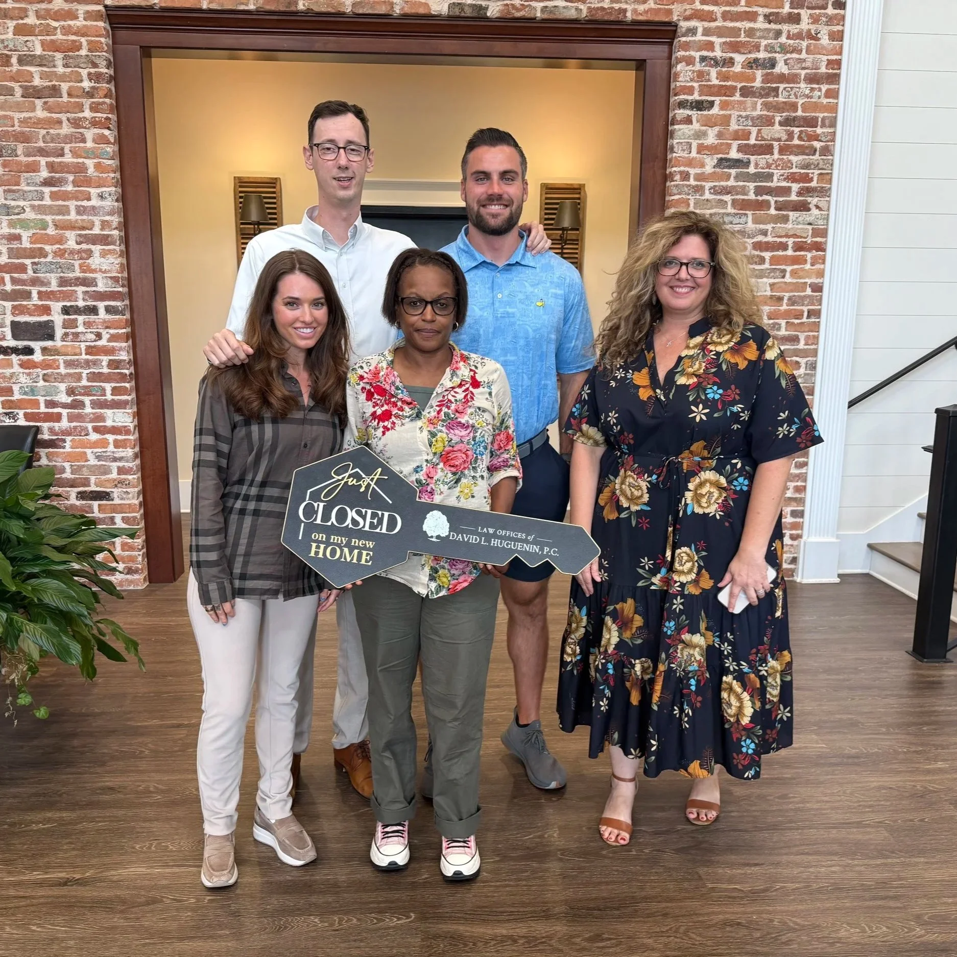 Group of five diverse adults smiling, standing indoors in front of a brick wall with a warm yellow background, holding a sign that says "Just Closed on my New Home."