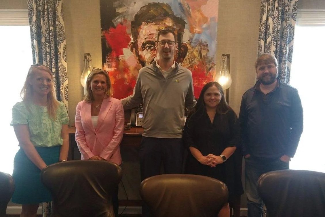 Group of five people standing in a conference room, with a colorful abstract portrait painting behind them.