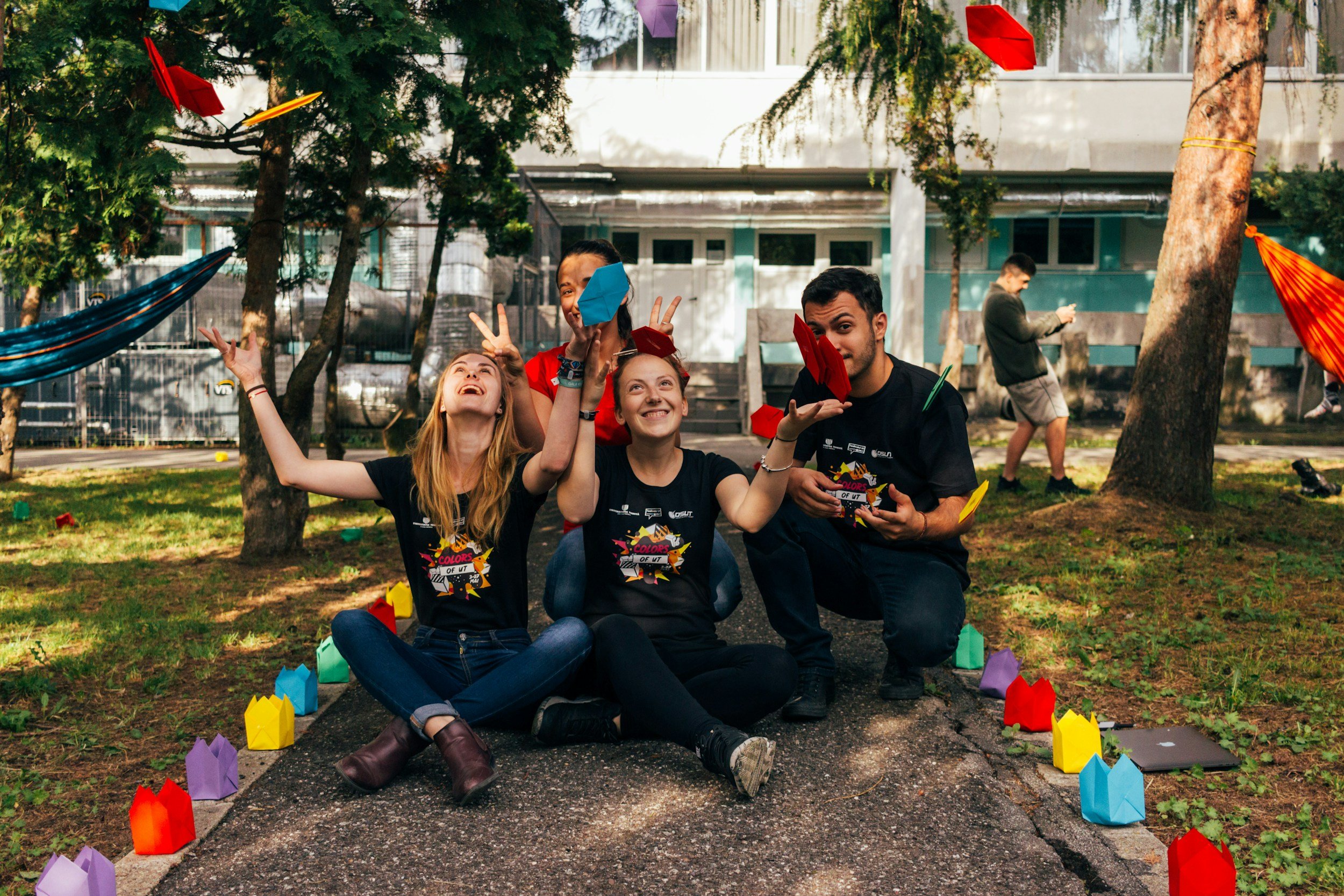Four young people sitting outdoors on a pathway surrounded by colorful paper lanterns, enjoying a celebration with paper airplanes flying around, with trees and a building in the background.