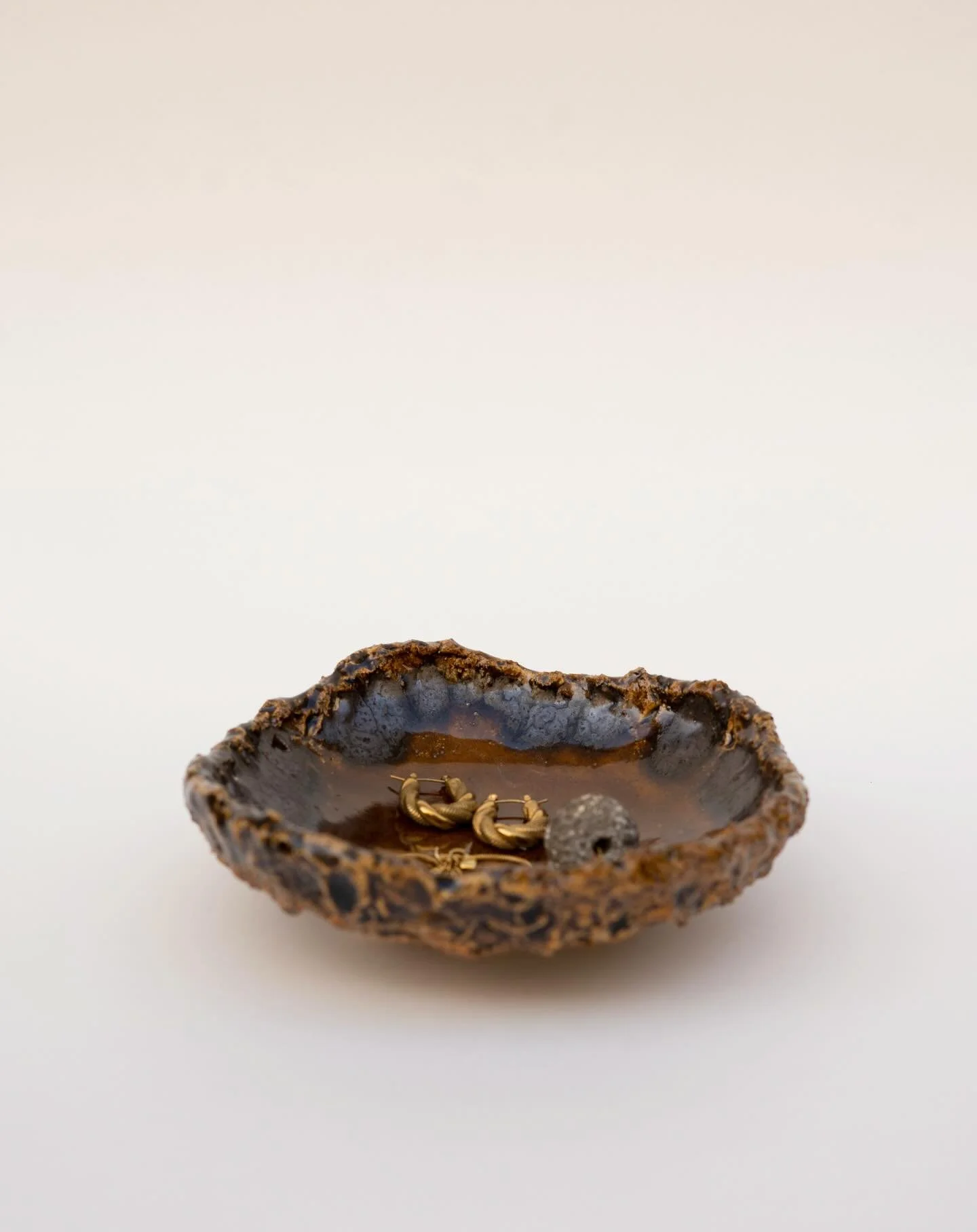 Handbuilt in stoneware, this trinket dish is made by Kelly Begiazi in Barcelona. Finished in a rich, dark honey glaze, the piece has an organic, irregular form with a textured rim. Designed to hold jewelry and small objects, it brings a grounded, dis