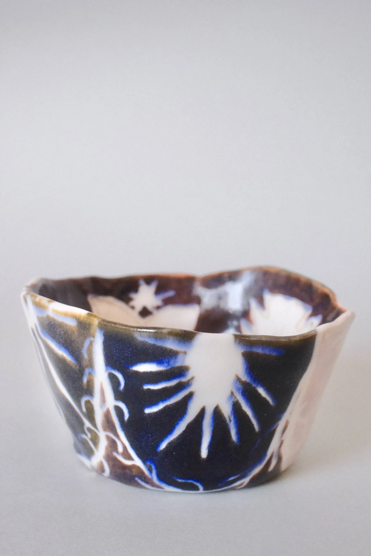 Hand-pinched porcelain cup with a flat base and irregular rim in brown, blue, pink, lavender and white