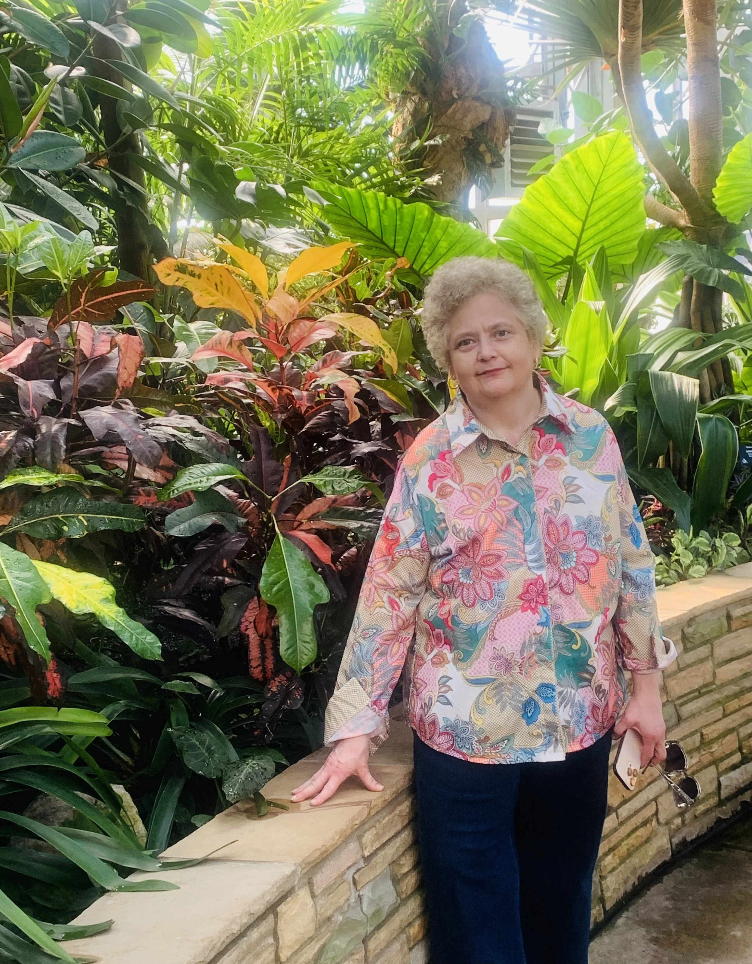 An older woman with curly gray hair standing by a brick wall surrounded by lush green and colorful tropical plants, holding sunglasses and a smartphone.