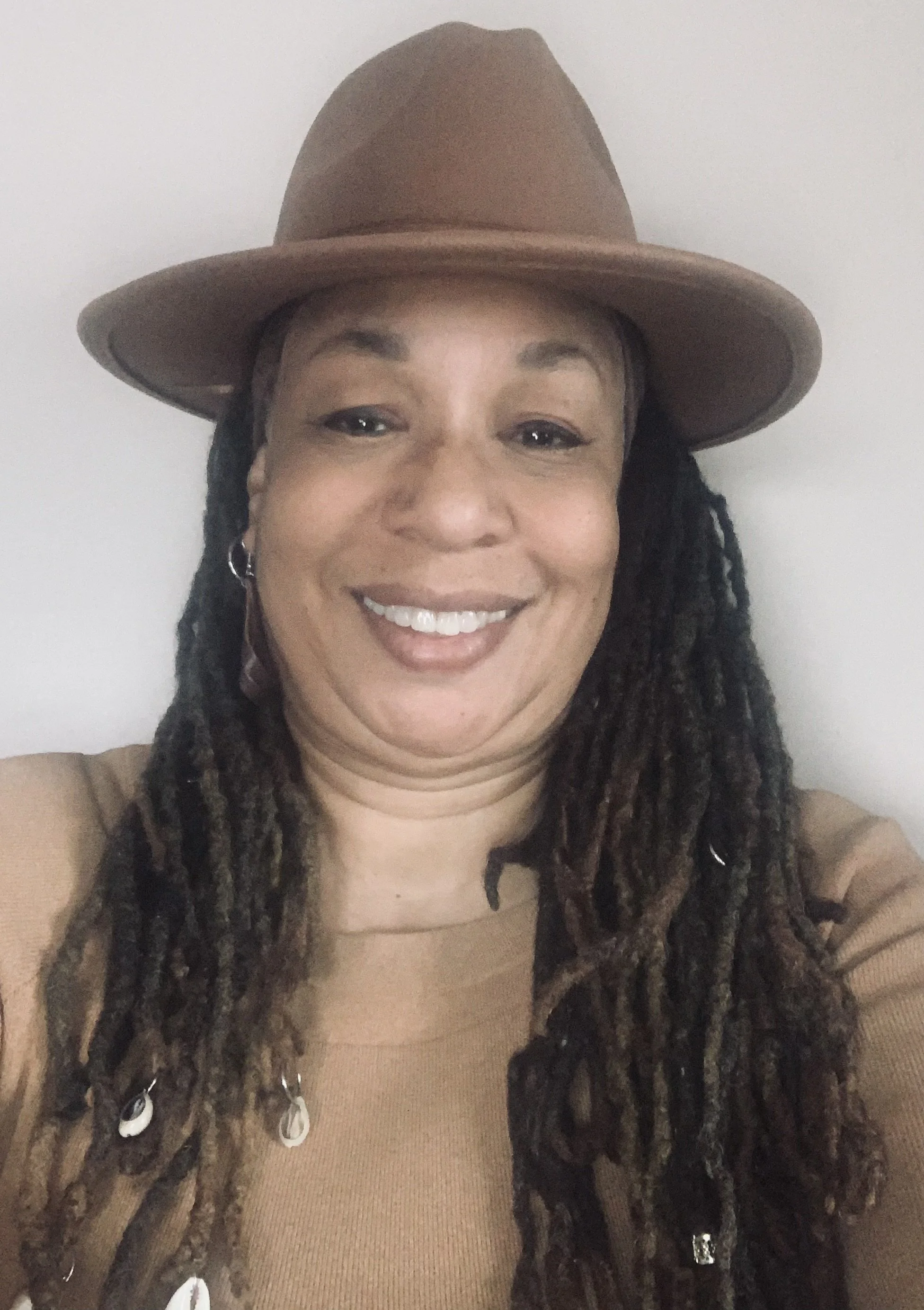 A woman with long dreadlocks wearing a brown hat and a brown shirt, smiling.